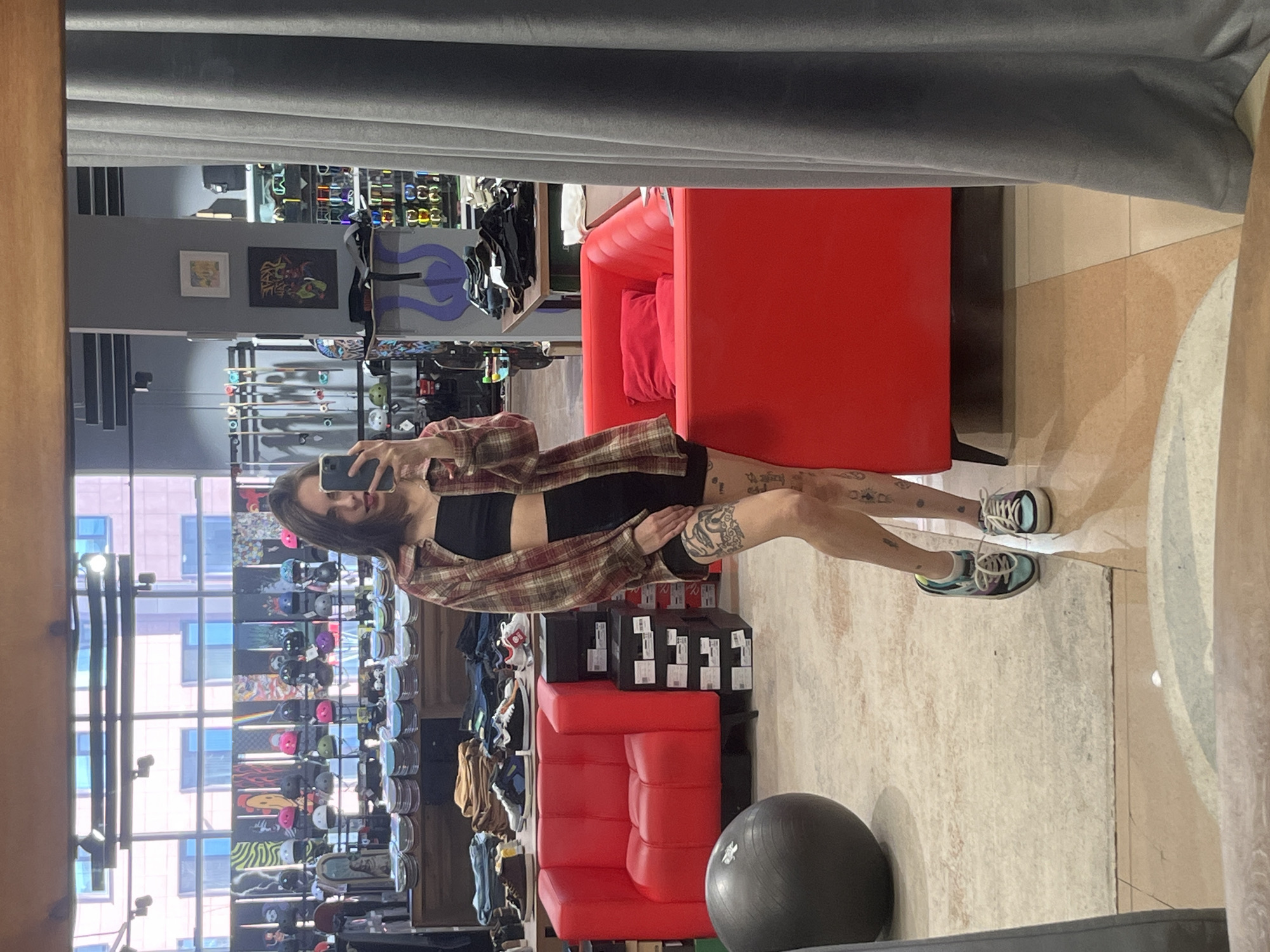 A mirror selfie captures a young woman standing within what appears to be a retail store in Krasnoyarsk, Russia. The woman, with long dark hair, looks towards her dark-colored smartphone, holding it up to take the photo. She is wearing a plaid button-down shirt open over a black crop top, black shorts, and multi-colored sneakers (light blue/green and black). Several tattoos are visible on her arms and legs.
The setting is an indoor space, likely a sports or skate shop, featuring display racks laden with skateboards, helmets, and other accessories in the background. To the right of the woman, a distinctive red modular couch or seating area is prominent. Above it on the wall, two framed pieces of art are visible. The scene is illuminated by natural light entering through large windows, which reflect urban buildings outside, indicating it is daytime. A large, dark grey exercise ball rests on the light-colored floor in the foreground. A grey curtain or fabric hangs overhead, and a wooden frame borders the left side of the mirror. No discernible text is clearly legible on any items or displays.