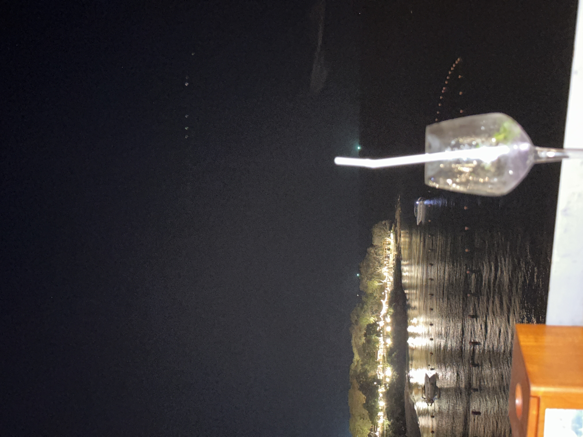 At night, a low-angle, close-up shot captures a scene in Pattaya City, Thailand. A clear glass holding a beverage and a white straw sits on an outdoor table, partially obscuring the background. The outdoor setting overlooks a body of water where lights reflect. Lights dot the shoreline where trees are. Several water vessels can be seen in the water.