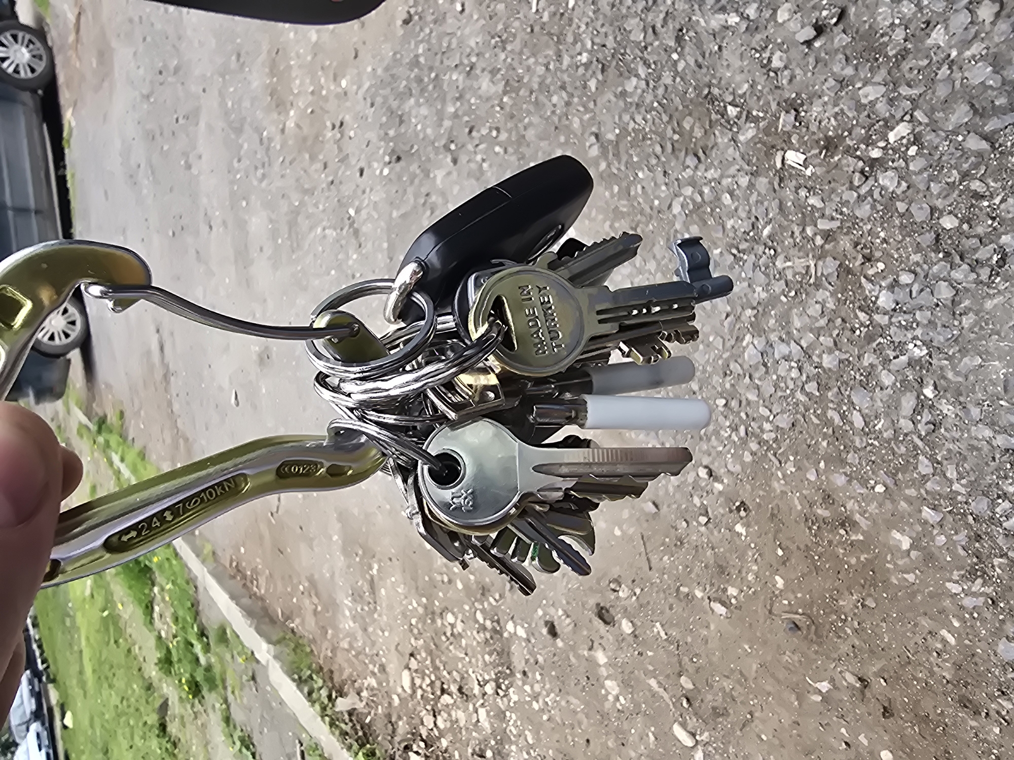 A hand holds a metal carabiner loaded with numerous keys on key rings. The keys include a car key fob and keys labeled "MADE IN TURKEY." The background shows a concrete surface with patches of grass and greenery. A partial view of a car is visible in the background. The precise location is impossible to determine from the provided image. There is no indication that this photograph was taken in Sofia, Bulgaria.