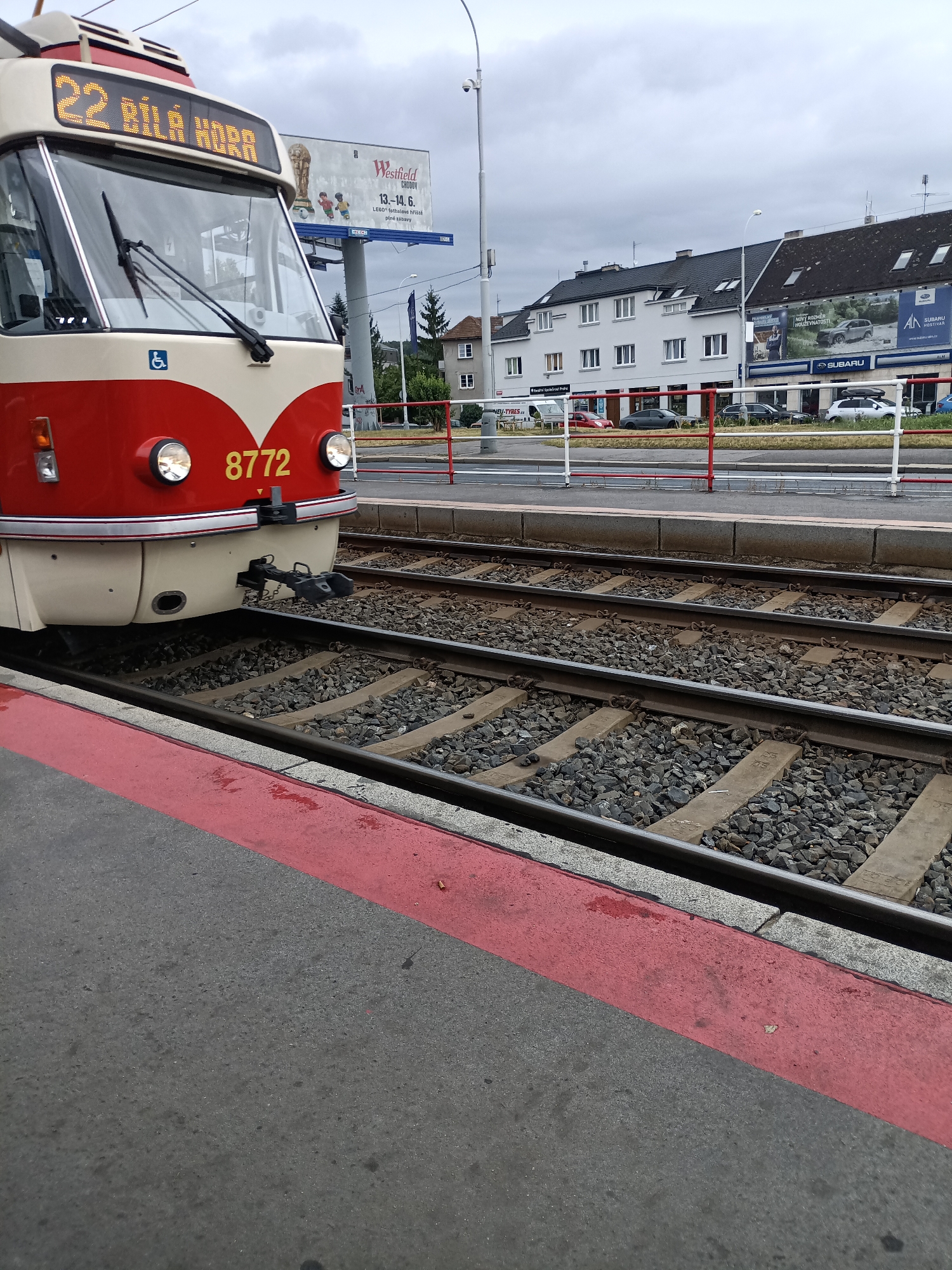 A low-angle, close-up shot shows the front of a red and cream colored tram numbered 8772, stopped at a tram station. The tram has a destination display above the windshield that reads "22 BÍLÁ HORA" in yellow LED text. A blue and white accessibility symbol is visible on the left side of the tram, below the windshield. Two round headlights are lit on the front of the tram. The tram is positioned on railway tracks with ballast.
In the background, across the tracks, a billboard features an advertisement for "Westfield Chodov" with a promotion for LEGO football figures from June 13-14. Further to the right, a row of buildings is visible, with signage for "Subaru Hostivice" on one of them. A white Subaru car is parked in front of the dealership. The sky is overcast, and the overall environment suggests an urban setting. A red and white striped barrier is visible in the midground.
