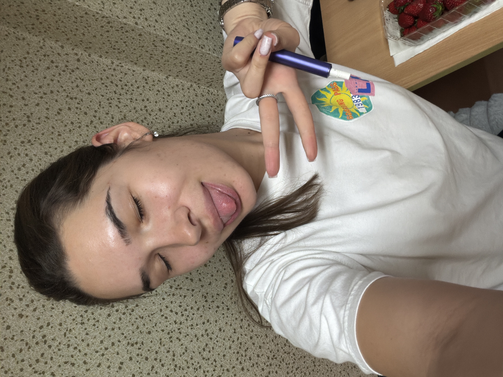 A young woman lies on a carpeted floor. She is wearing a white t-shirt with a graphic design that includes a sun and the text "Sunshine Pass 1983." She is holding a purple electronic cigarette in her hand and making a peace sign with her fingers. Her eyes are closed, and she is sticking her tongue out. To the right of her, a table holds a plastic container of strawberries. The location is presumed to be in Sofia, Bulgaria, based on the query context.