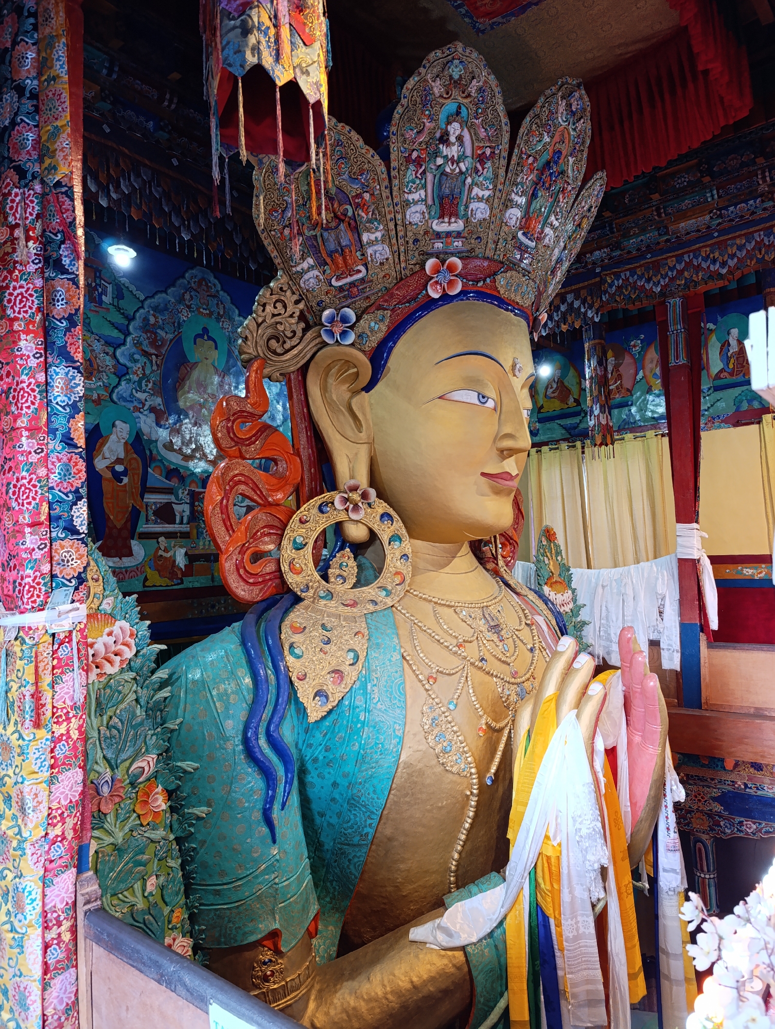 The image shows a large, gold-colored statue of a deity within a temple. The statue is adorned with an ornate, jeweled crown and elaborate necklaces. It is depicted with its hands pressed together in a gesture of prayer or blessing, with colorful prayer flags draped around them. The background features vibrant murals and colorful tapestries depicting religious scenes and figures. The overall environment suggests a Buddhist or Hindu place of worship.
