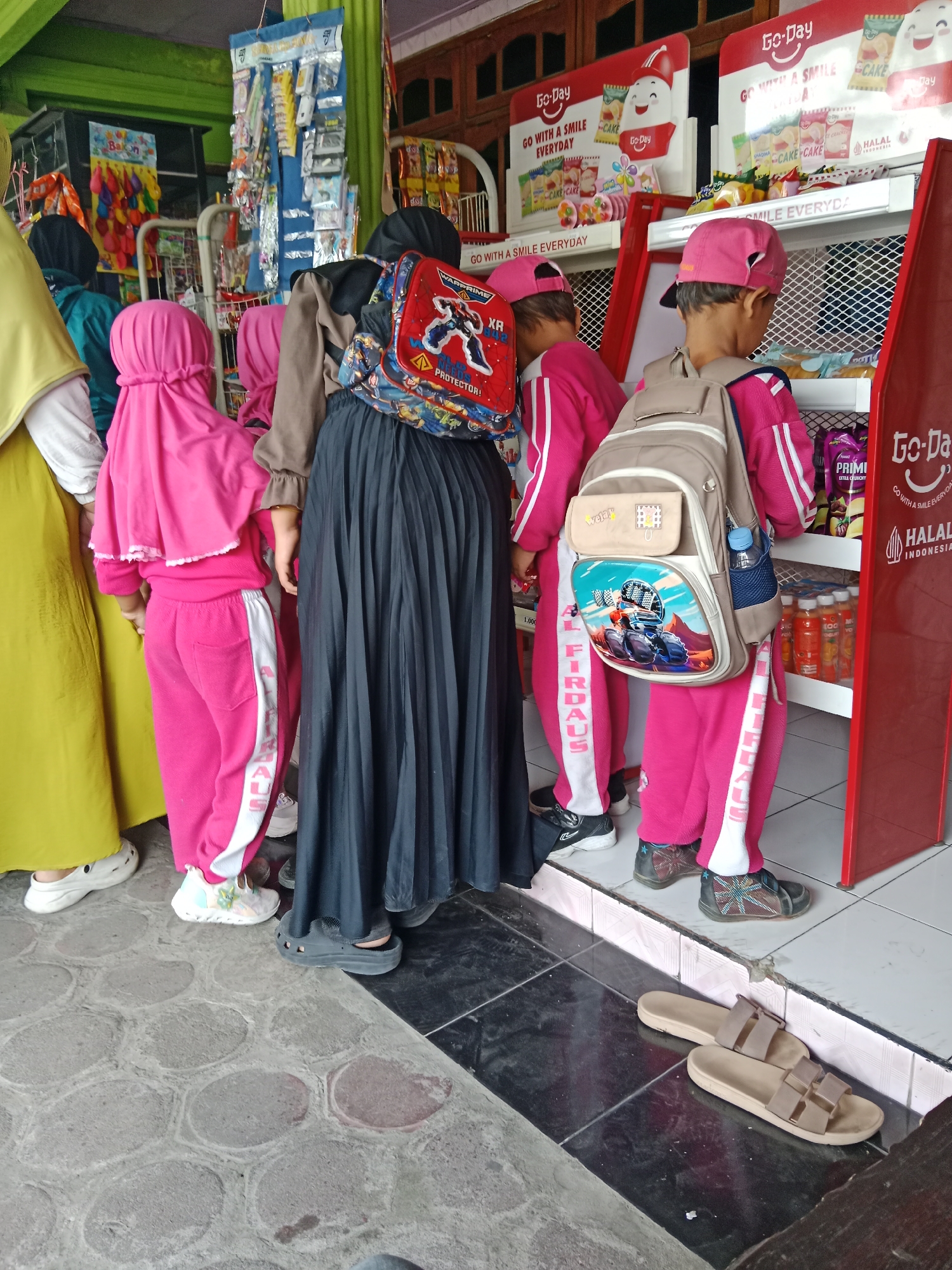 The image depicts the exterior of a small convenience store, likely a warung, in Sambi, Indonesia. The store features a red and white facade, with shelves stocked with various packaged goods, including snacks and drinks. Two young children, dressed in matching pink tracksuits and headscarves, are standing near the shelves, possibly shopping with an adult. The adult, wearing a long black pleated skirt and a brown hijab, is partially visible. Another person in a yellow hijab is also present. The ground is paved with patterned tiles. The overall scene suggests a typical day at a local Indonesian shop.