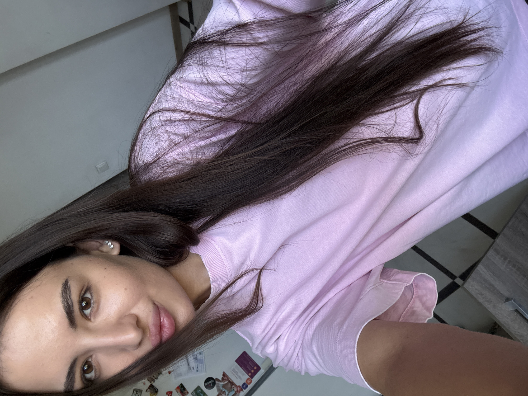 A young woman with long, dark hair and brown eyes is taking a selfie in a well-lit room. She is wearing a light pink t-shirt. Her left ear has a small, diamond-like earring. The background shows a white wall with some light fixtures and a dresser with a patterned surface. There are also some items on the wall to the left, which appear to be magnets on a refrigerator. The overall setting suggests a residential interior, possibly in an apartment or house in Chișinău, Moldova. The lighting is natural, coming from a window or overhead source, casting soft shadows.