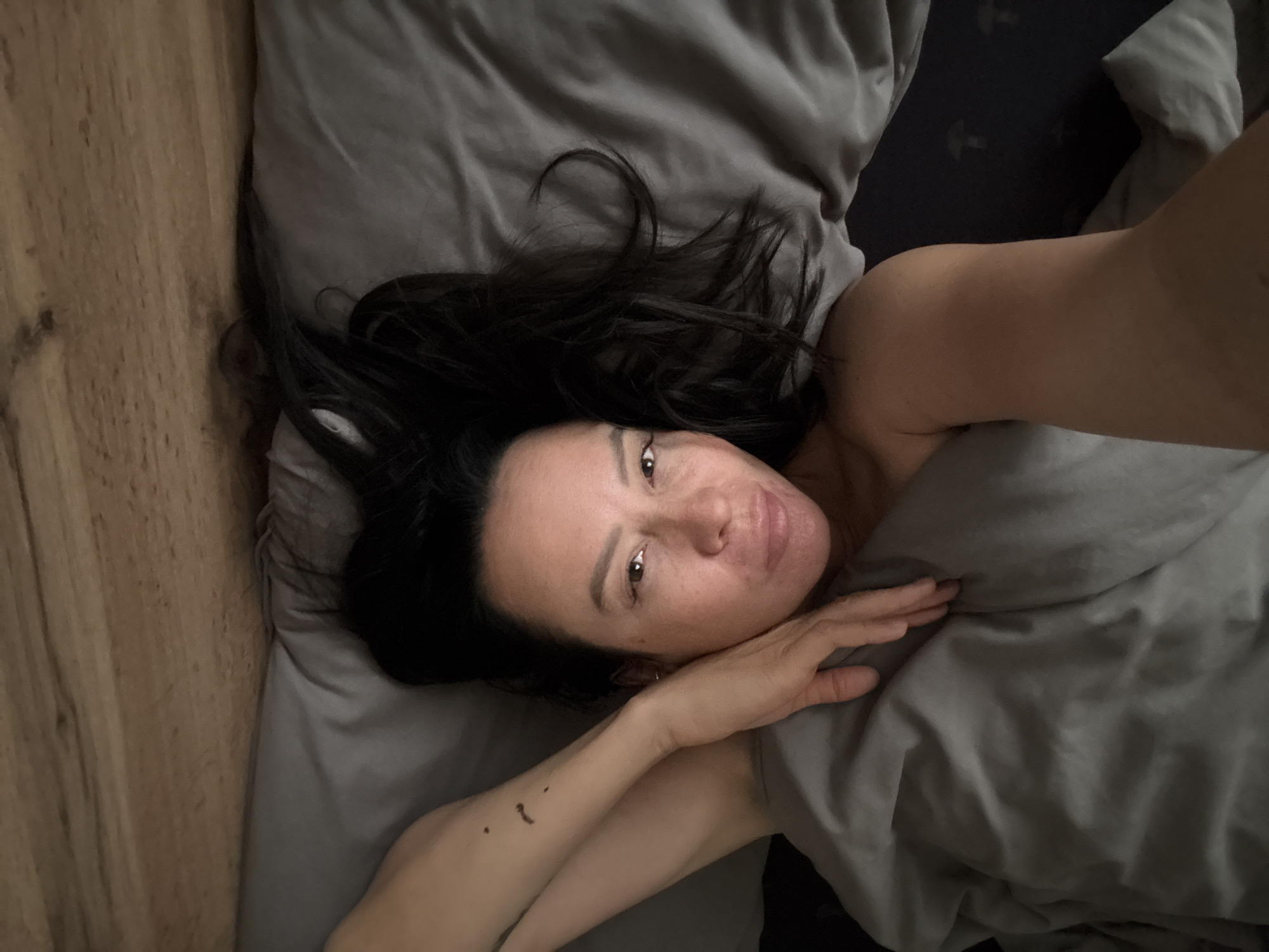 A woman with dark hair lies on a gray bed with a gray duvet. She is resting her head on the pillow. Part of a wooden floor is visible on the left side of the frame. The location is presumed to be Chaikovsky, Russia.