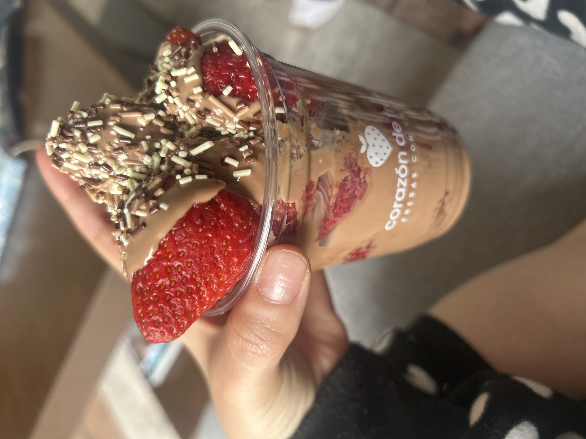 A person is holding a clear plastic cup filled with strawberries and chocolate sauce, topped with chocolate sprinkles and white sprinkles. The cup has a logo that reads "corazón de fresas con". The person is wearing a black shirt with white polka dots. The background is blurred, but it appears to be a casual setting.