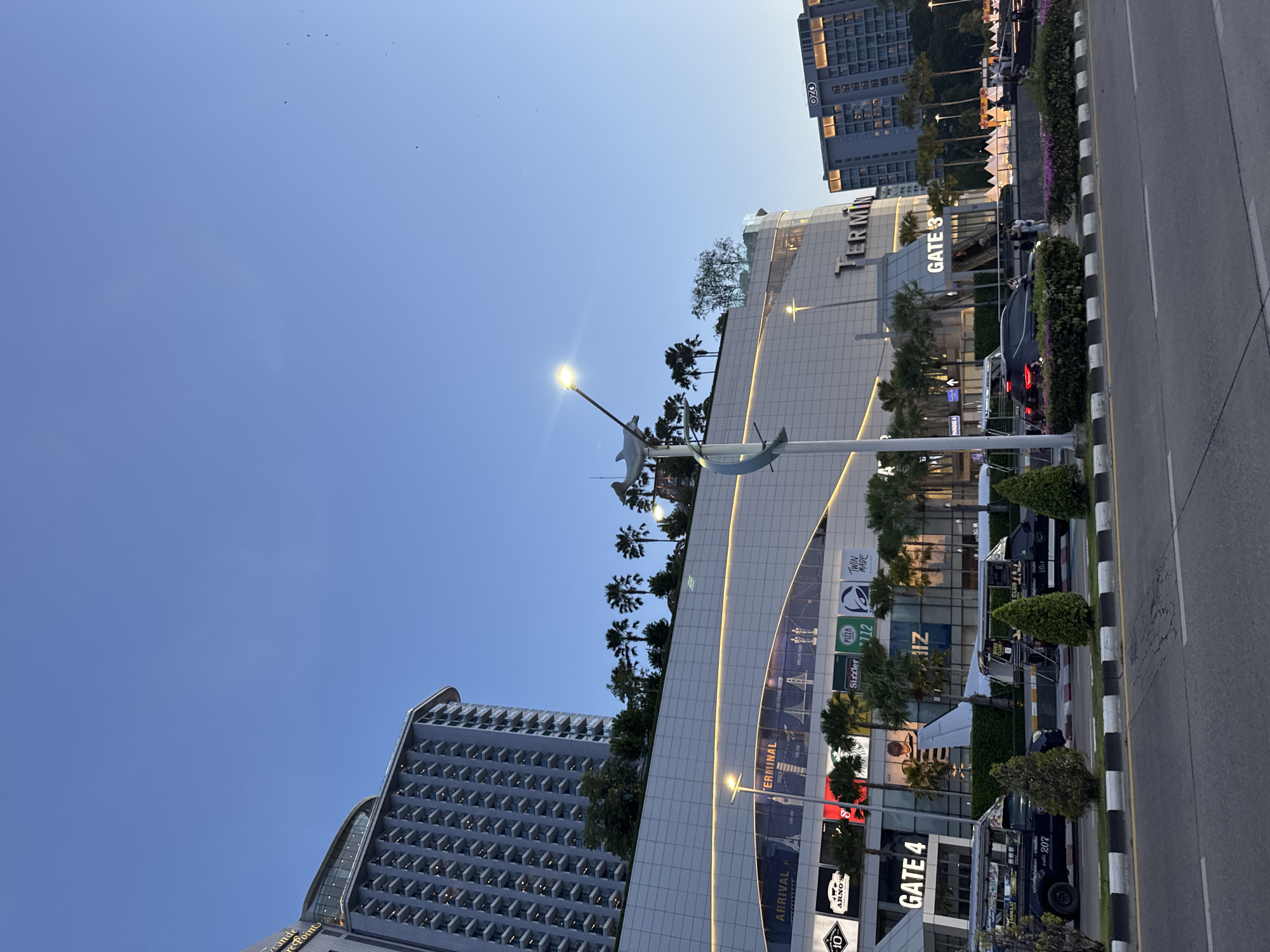 This is a photograph of a large, modern shopping mall complex in Pattaya City, Thailand, taken at dusk. The mall, labeled "TERMINAL," has a multi-story facade with large glass windows and illuminated signage. Several well-known international brands are advertised on the facade, including Starbucks, Sizzler, and Taco Bell. To the left of the mall is a tall, high-rise hotel building with a distinctive architectural style, featuring numerous balconies and a curved top. In front of the mall, a wide paved road is visible with cars and a few pedestrians. The road is bordered by a sidewalk and a landscaped area with palm trees and flowering bushes. The sky is clear and blue, indicating the end of the day. The overall scene depicts a bustling urban environment with a mix of commercial and residential structures.