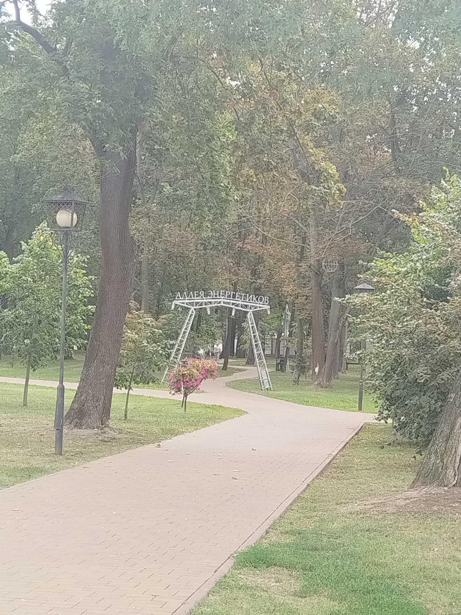 In the city of Brest, Belarus, a paved park pathway curves through a verdant, tree-lined area. A lamppost stands on the left, and an archway with the inscription "АЛЛЕЯ ЭНЕРГЕТИКОВ" (Alley of Energetics) is visible in the background. The archway is a metallic structure with diagonal supports and lights. Pink flowers bloom near the base of the arch. The grass is green, and the trees exhibit foliage of various shades of green and brown, suggesting it might be late summer or early autumn. Several people are walking in the distance near the archway. The overall scene depicts a public park setting.