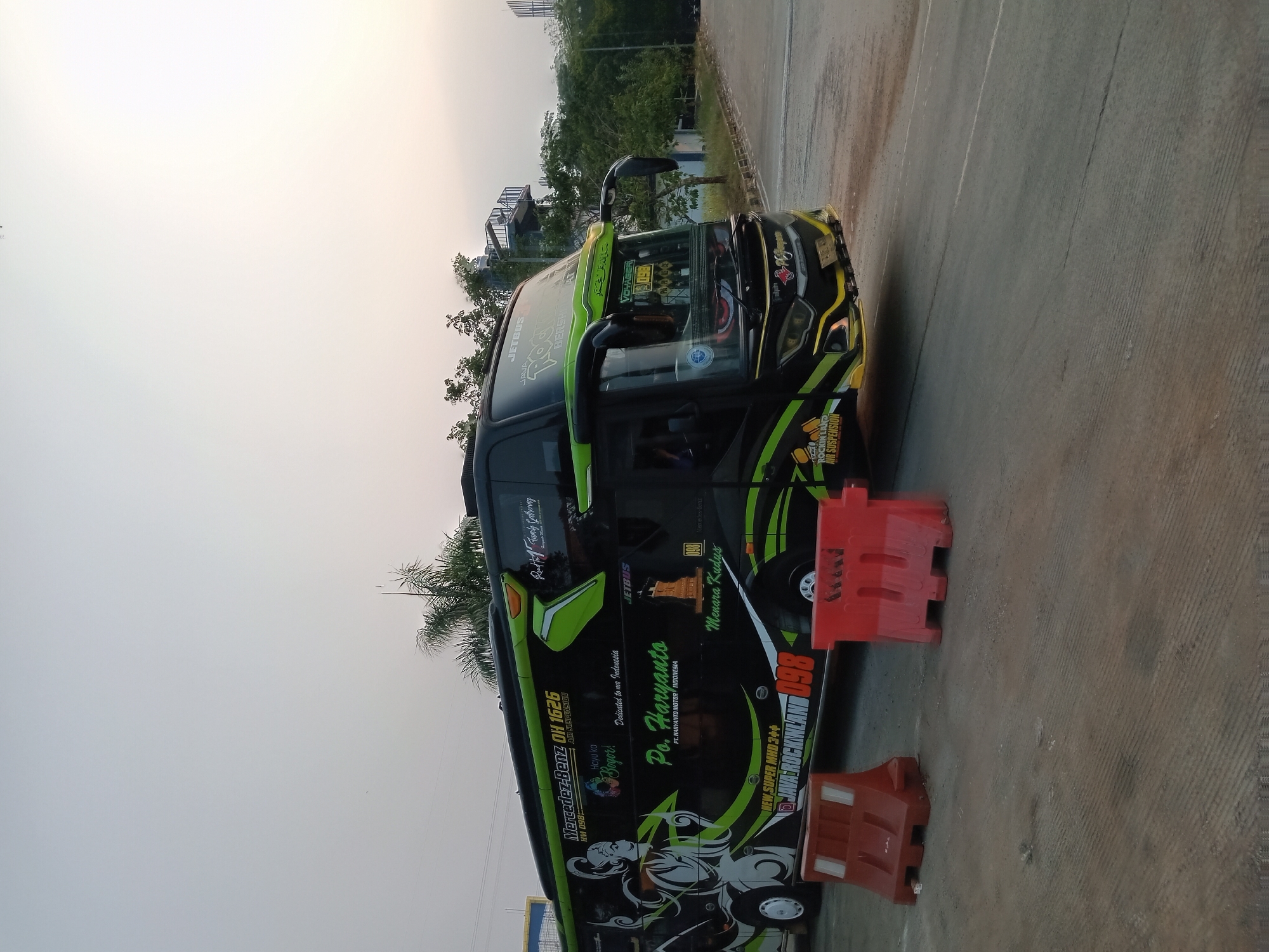 The image shows a large, black and green intercity bus parked on a paved surface with two orange traffic barriers in the foreground. The bus has several decals and writings on its side, including "Po. Haryanto," "Mercedes-Benz OH 1626," and "Menara Kudus." The bus also features intricate graphic designs. In the background, there are trees, a building with an elevated walkway or structure, and what appears to be a sky with a hazy or overcast quality, suggesting it may be early morning or late afternoon. The scene suggests the bus is at a rest stop, terminal, or parking area. The location is identified as Depok, Indonesia.