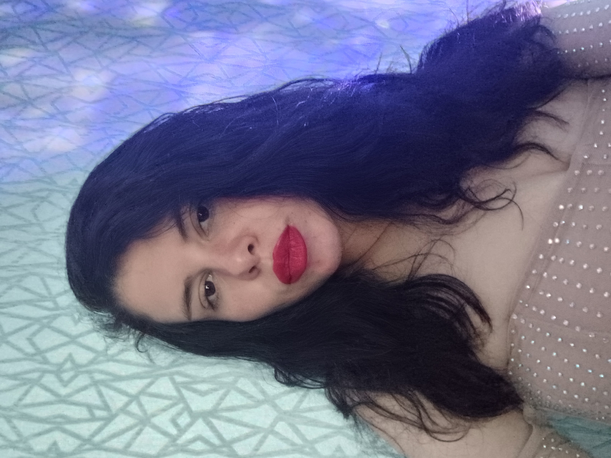 A woman with dark hair and fair skin is lying on a bed. She is wearing a beige dress with small, reflective embellishments. Her lips are painted red. The bed has a geometric pattern in light blue and white. The lighting is soft, with some blue hues, and there is no evidence of a location in Sofia, Bulgaria.