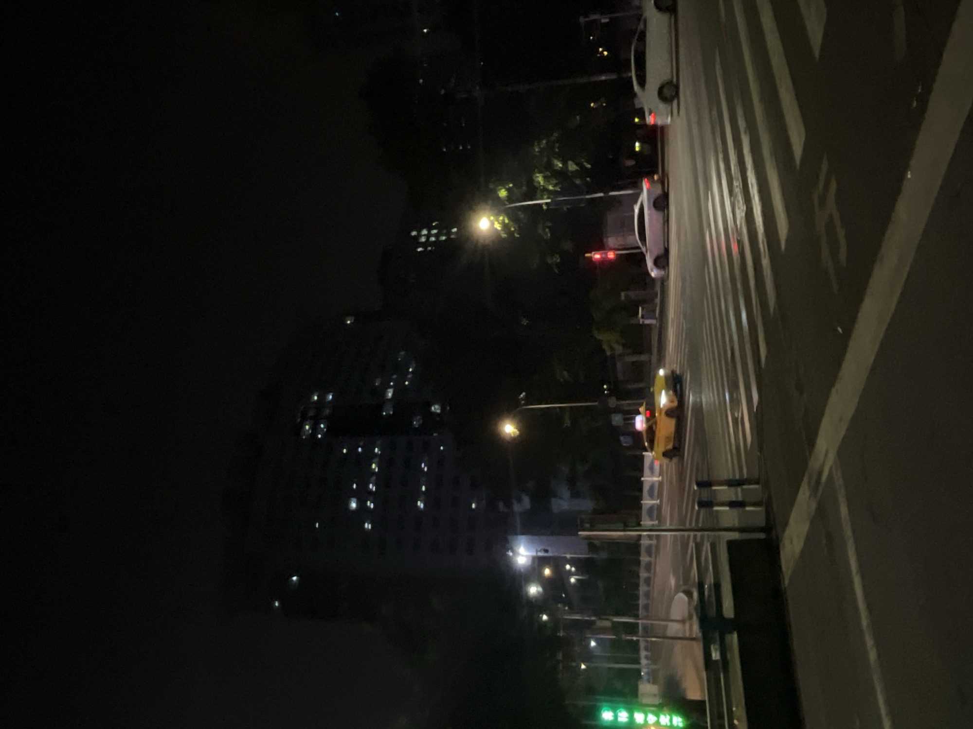 The image depicts a street scene at night in Yuzhong District, China. A yellow taxi is turning right at a crosswalk, with its headlights on. Two white cars are visible at the intersection, one in the left lane and one in the right lane. A tall, dark building with many illuminated windows dominates the background. Streetlights illuminate the intersection and surrounding trees. The road surface shows reflections from the lights, indicating it may be wet. A pedestrian crossing is clearly marked with white stripes. A traffic light shows a red signal for the lane the white car is in, while the taxi appears to be proceeding. Green traffic signals are visible on the left side of the frame.