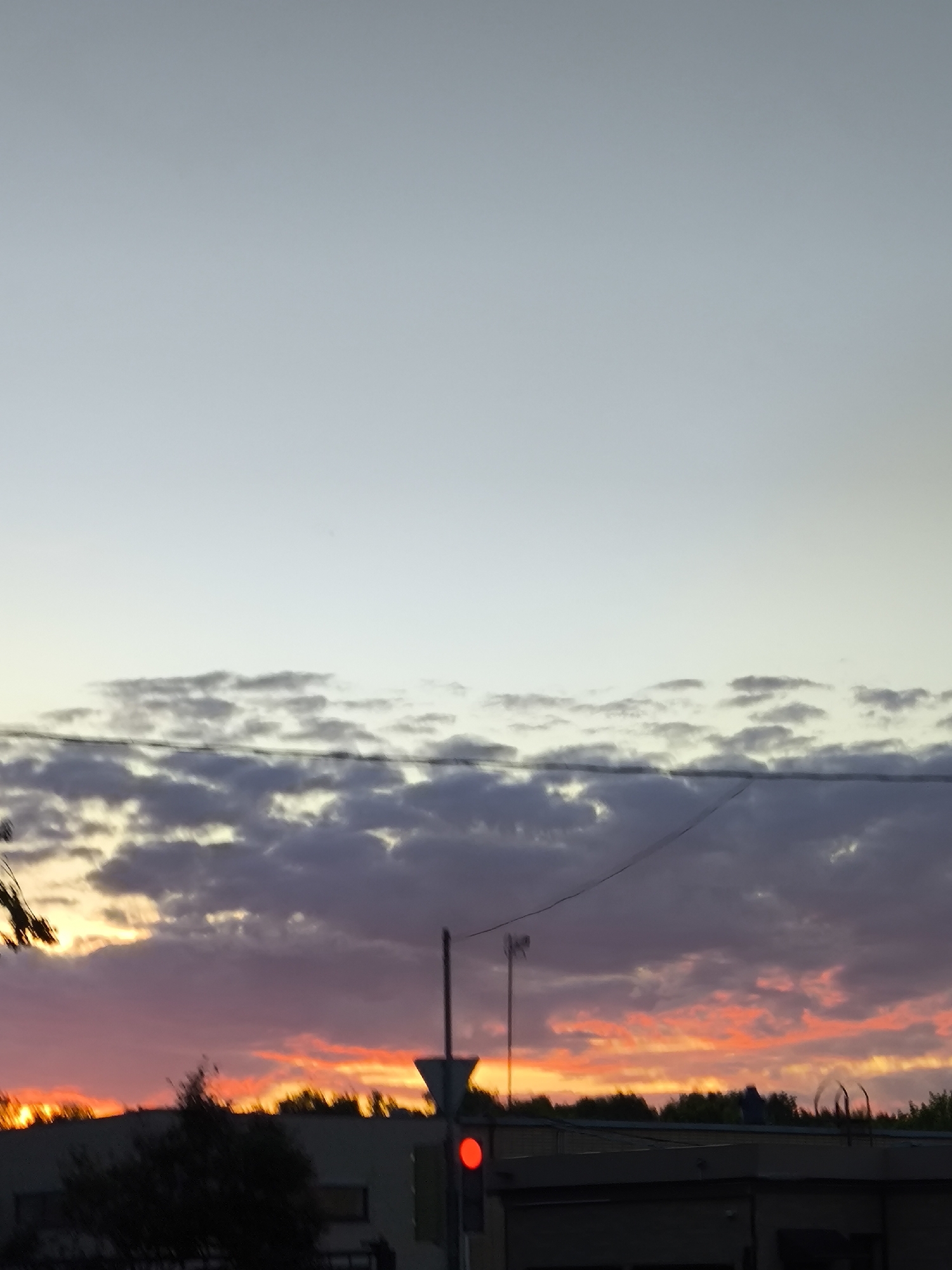 The image displays a vibrant sunset over an urban or suburban landscape. The sky dominates the frame, showcasing a gradient of colors. The uppermost section is a pale blue-grey, transitioning downwards to soft purples and greys where scattered clouds are present, and finally erupting into intense oranges and reds along the horizon, indicative of the setting sun.
A band of cumulus clouds stretches across the mid-section of the sky, appearing darker where they obscure the sun's direct light and brighter with warm hues below. Power lines or utility cables stretch horizontally across the middle of the sky.
In the lower third of the image, the silhouettes of buildings and trees are visible against the bright sunset. A tall, slender pole extends upwards from the ground on the right, near which a traffic light is clearly visible, illuminated red. Additional dark, silhouetted foliage is present on the far left, suggesting trees or bushes. The scene conveys the late hours of the day, with the dramatic colors of dusk illuminating the sky above urban structures. There is no visible text.