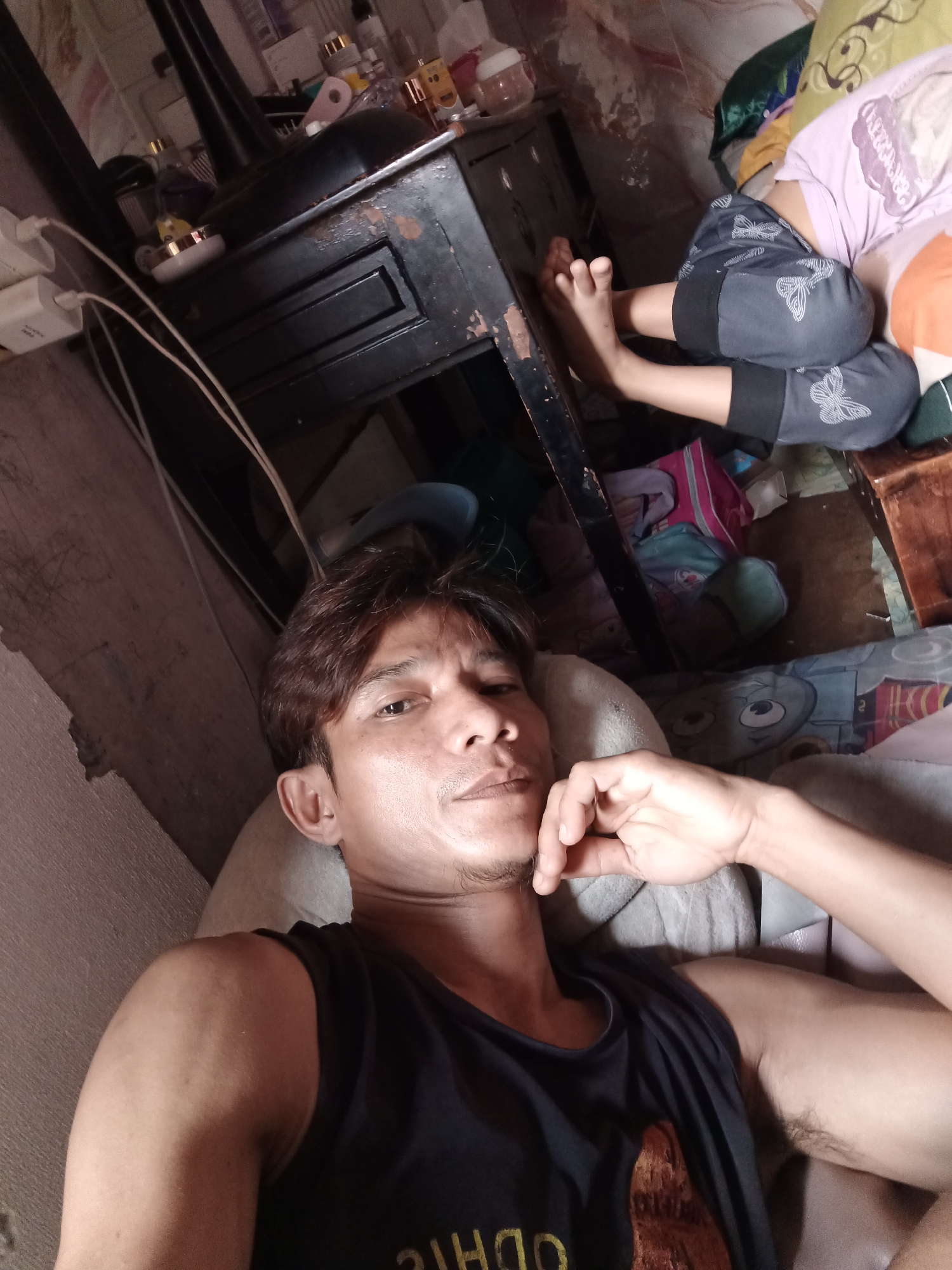 The image shows a man lying down in what appears to be a bedroom. He is wearing a black tank top and his right arm is raised, touching his face. In the background, a child is seen lying down, wearing a purple t-shirt and grey shorts with butterfly patterns. There are various items scattered around, including bags, bottles, and electrical chargers. The room appears cluttered and somewhat dishevelled, with visible wires and a wooden dresser. The lighting suggests it is indoors, possibly during the day with artificial light also present. No specific landmarks of South Jakarta are identifiable in the image.