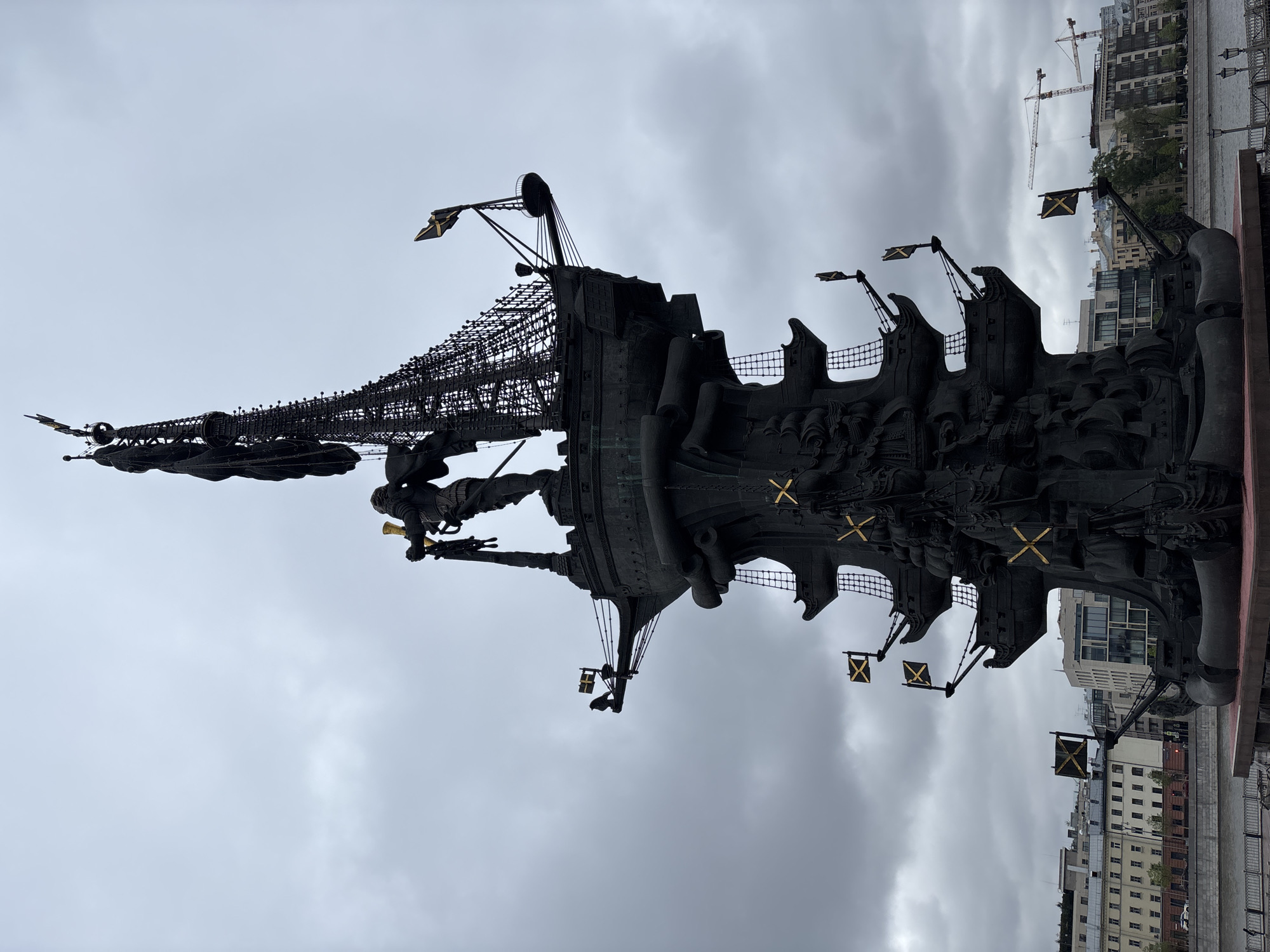 The image depicts the Peter the Great Statue in Moscow, Russia. The statue is a tall monument of Peter the Great standing on a ship, with flags bearing a cross symbol hanging from the ship's masts. The statue is made of dark metal and is set against a cloudy sky. Buildings and cranes are visible in the background.