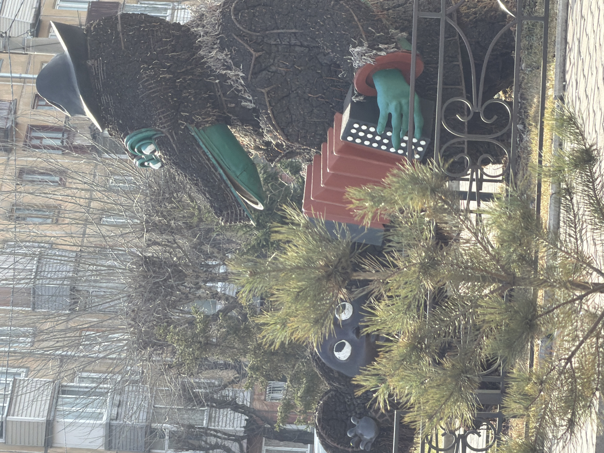 The image captures a whimsical outdoor scene in Krasnoyarsk, Russia, depicting a decorative figure resembling a gnome or forest creature, constructed from dark, textured material that mimics tree bark or roots. The figure is positioned to appear as if playing an accordion.
The main subject is the upright figure, which features a wide-brimmed, black, pointy hat at the top. Two bright green, gloved hands emerge from the figure; one hand is actively positioned on the black and white keys of a reddish-brown accordion. The accordion has distinct red-orange bellows. Towards the bottom of the figure, a dark face with large, round white eyes is visible, peeking out from behind green pine needles. Another green element, possibly a boot or part of clothing, is also visible.
In the foreground and middle ground, vibrant green pine branches and needles partially obscure the figure. A black wrought iron fence with decorative scrolls runs vertically in the center-right. The background consists of several multi-story apartment buildings, characteristic of Soviet-era architecture, with numerous windows. Many bare tree branches, suggesting a late autumn, winter, or early spring season, are prominent in front of these buildings, further hinting at the time of year.
The scene is brightly lit by natural sunlight, indicating daytime with clear weather. There is no visible text in the image. The overall impression is that of a charming, possibly artistic, public installation situated within an urban residential setting.