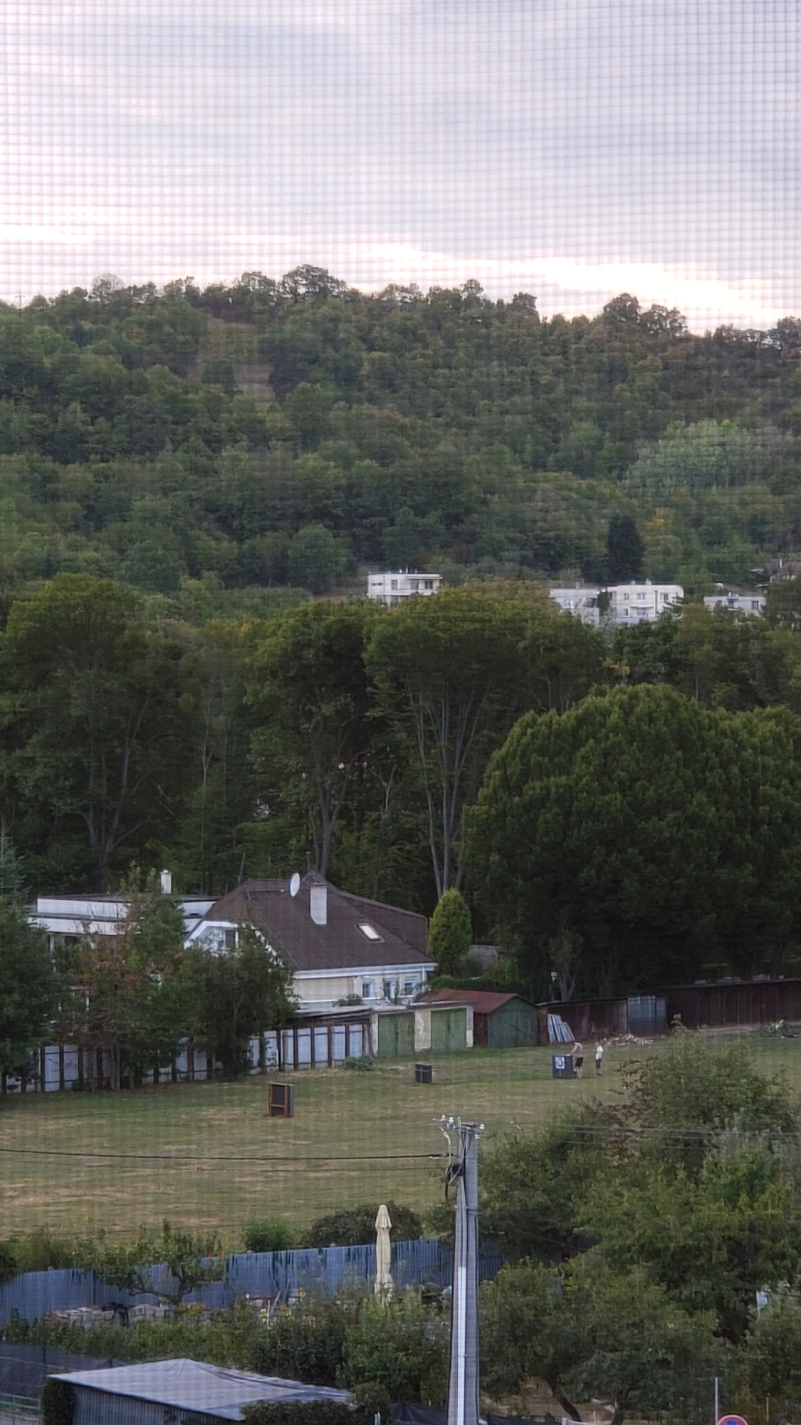 The image depicts a panoramic view of a green hillside covered with dense trees, suggesting a suburban or semi-rural setting on the outskirts of Prešov, Slovakia. In the foreground, a grassy field is visible, with several structures including a house with a brown roof and white walls, two small sheds, and a fence. Further back, amidst the trees, several white apartment buildings are situated on the slope of the hill. Two individuals are standing in the field near a blue target, indicating a recreational activity such as archery or shooting. The sky above is overcast with a pale, diffused light, hinting at a possibly late afternoon or early morning scene. The overall impression is one of a peaceful, natural landscape integrated with residential buildings.