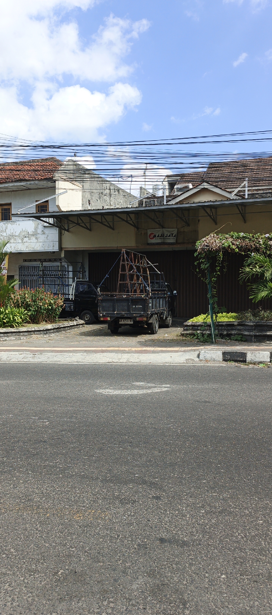 The image depicts a street scene in the city of Yogyakarta, Indonesia, under a partly cloudy sky. In the foreground, a gray asphalt road occupies the bottom half of the frame. A white and black striped curb runs along the edge of the road. Behind the curb, a narrow strip of dry grass and some sparse vegetation is visible.
The mid-ground of the image features a row of buildings with awnings extending over the sidewalks. The buildings have tiled roofs in various shades of red and brown. One building has a sign that reads "MAJU JAYA." In front of the buildings, two black pickup trucks are parked. The truck in the foreground has a metal rack structure in its bed. The license plate on this truck is partially obscured but appears to read "AB 8910 KH."
The background shows more buildings and a clear blue sky with scattered white clouds. Electrical wires are strung across the top of the frame, casting shadows on the scene below. The overall impression is a typical urban street view in Indonesia, possibly during daylight hours given the lighting conditions. There are no people visible in the image.
