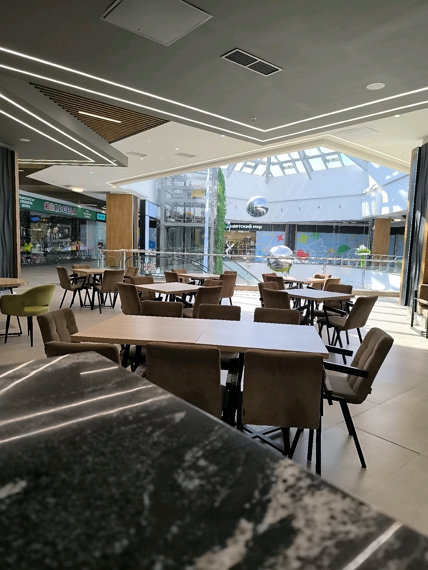 The image depicts the interior of a modern, well-lit shopping mall food court. The foreground is dominated by a large, dark, speckled table, likely made of granite or a similar stone. Beyond this table, numerous dining tables and chairs are arranged in rows. The tables have light-colored wooden tops and dark metal frames. The chairs are upholstered in a neutral, taupe fabric and feature dark metal legs. In the mid-ground, a variety of shops are visible through large glass windows, including one with a prominent sign reading "ДЕТСКИЙ МИР" (Children's World). An escalator and an elevator are also visible in the background, suggesting multiple levels of the mall. The ceiling features a complex design with recessed lighting and geometric panels. A large, reflective spherical object, possibly a disco ball or decorative element, hangs from the ceiling in the central atrium area, which is topped with a large skylight allowing natural light to flood the space. The overall impression is one of a clean, contemporary, and spacious public area, characteristic of a modern shopping center. The provided information about the location in Qarağandı city administration, Kazakhstan, places this scene within a specific urban context. There are no people visibly present in the image.
