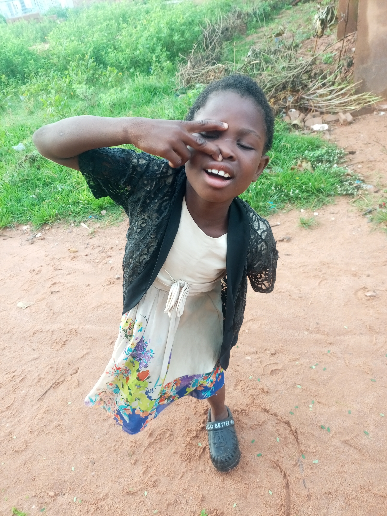 A young Nigerian girl stands outdoors in what appears to be a sandy, unpaved area in Tempure, Nigeria. She is wearing a light-colored dress with a floral print and a black lace overlay jacket. Her right hand is raised, with her fingers touching her nose and eye. She has her mouth open, showing her teeth, and her eyes are partially closed. Her left arm is not visible beyond the sleeve of her jacket, suggesting it may be amputated or held in a way that it is not discernible. She is wearing a black clog-style shoe on her visible right foot, with white lettering that reads "GO BETTER". The background consists of dry dirt, sparse green vegetation, and some dry brush. The lighting suggests it is daytime.