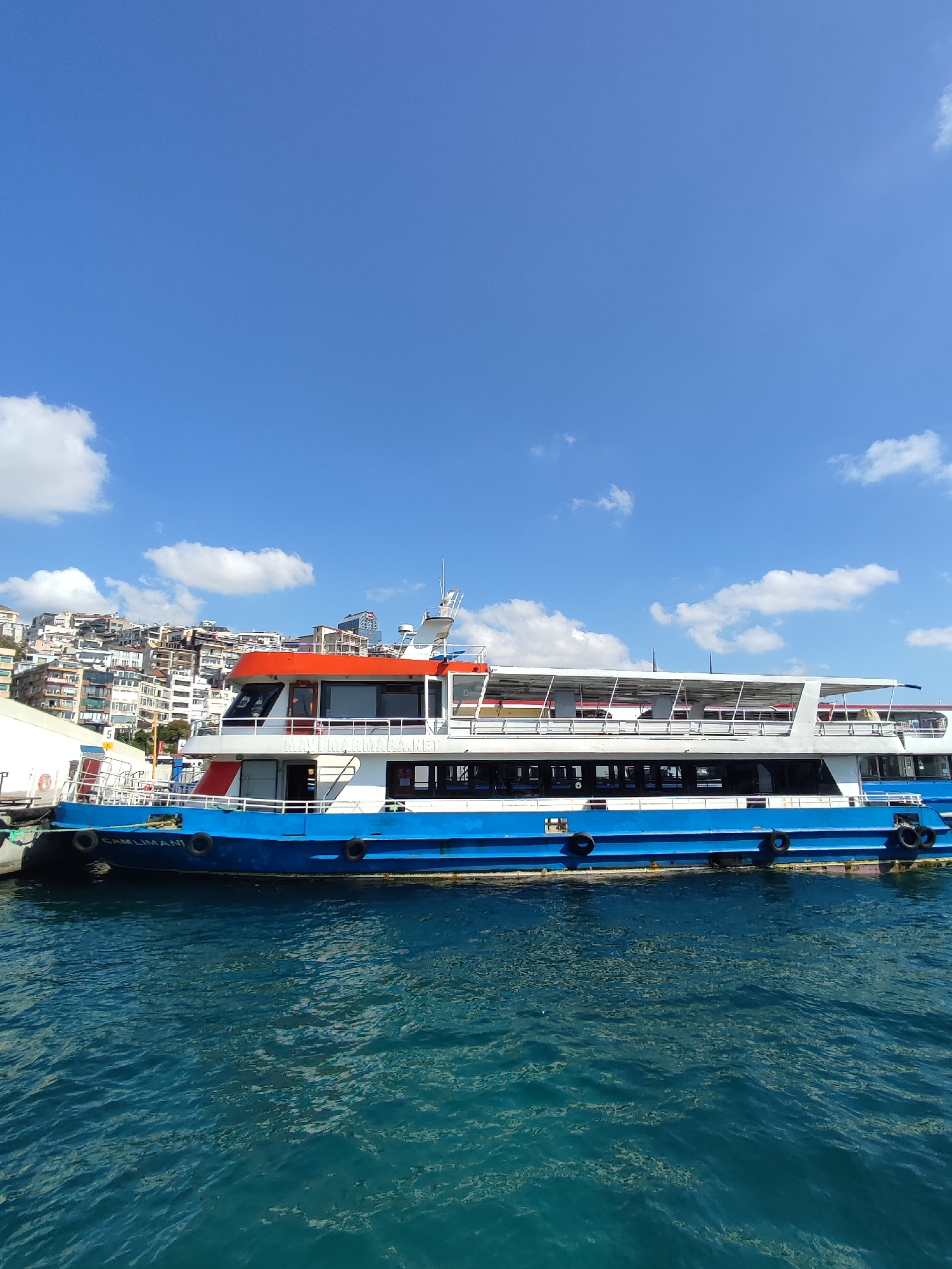 A blue and white ferry with an orange top deck is docked in the water. Buildings are visible on a hill in the background, with a clear blue sky and white clouds above. The ferry has multiple decks and windows, with the name "CAMLIMANI" visible on its side. The water is a deep blue and appears slightly rippled. The scene is bright and sunny. This scene is likely located near the Beyoğlu district of Istanbul, Turkey, as suggested by the presence of a ferry and the characteristic architecture of the buildings on the hillside.