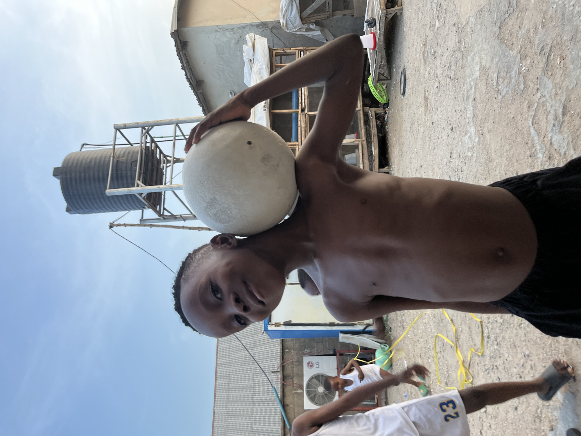 The image shows a shirtless child balancing a white ball on their head in Madalla, Nigeria. The child is dark-skinned and wearing dark shorts. The background shows a water tank on a metal frame, various buildings, and at least two other people. One person is wearing a white tank top. An LG air conditioner is visible on the side of a building. A yellow rope is on the ground. The ground appears to be dirt or concrete.