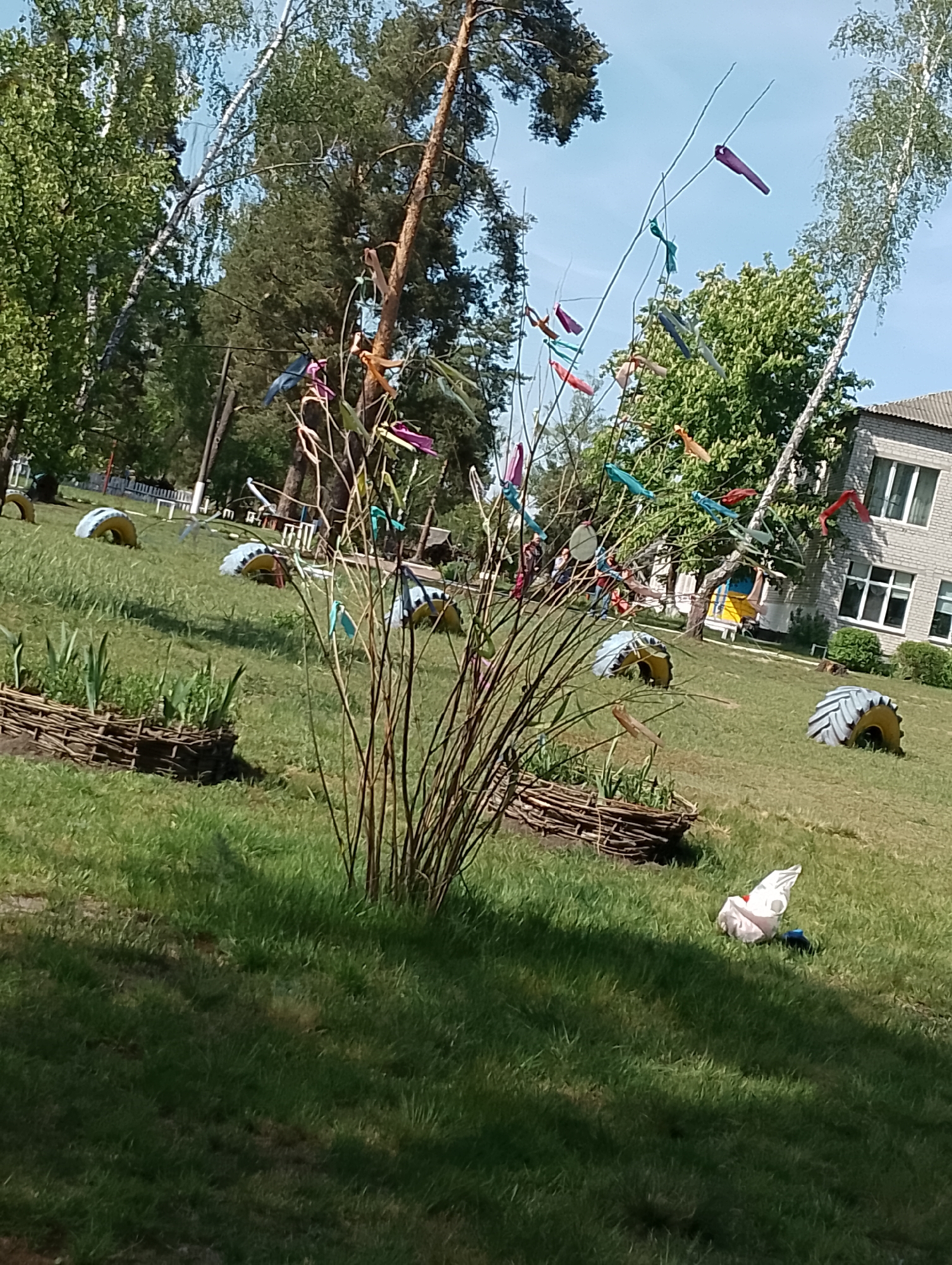 The photograph depicts a grassy lawn in an unidentified location. The lawn contains several repurposed tires and raised garden beds constructed of woven twigs, as well as a decorative bush with colorful ribbons attached. A white plastic bag is visible on the grass, and a two-story building with a gray roof and multi-paned windows stands in the background. Trees are visible throughout the scene, creating a partial canopy overhead.
