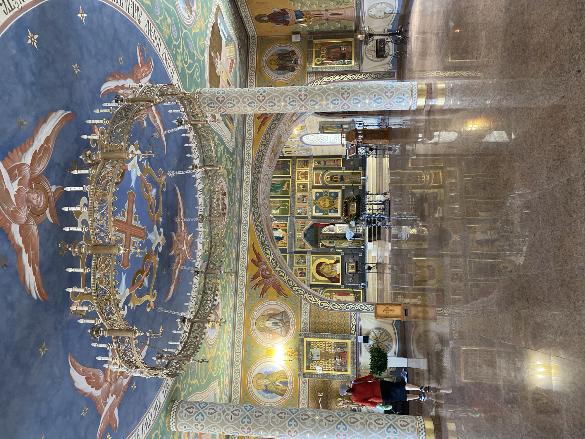 The image captures the opulent and richly decorated interior of an Orthodox church, identified by context as being in Nizhny Novgorod, Russia. The perspective looks upwards towards the high, vaulted ceiling and across a highly reflective floor.
The ceiling and upper walls are adorned with elaborate frescoes and iconography. A prominent central section of the ceiling is painted in a deep blue, featuring stylized angels with large, feathered wings, golden stars, and a large red cross. Hanging from this blue expanse is a large, ornate golden chandelier designed to hold numerous candles. Surrounding the blue section, other areas of the ceiling and walls display intricate patterns in shades of green, gold, and earthy tones, alongside circular medallions depicting saintly figures and other religious imagery.
Two substantial, cylindrical columns are visible, supporting the architectural structure. They are covered in intricate, repeating patterns of heart-like shapes, crosses, and foliage, rendered in blue, green, white, and gold.
The lower sections of the walls are covered with numerous icons, both large and small, many framed in gold, depicting various saints and biblical scenes.
The floor is a highly polished, reddish-brown marble or stone, creating striking, mirror-like reflections of the chandeliers, columns, and parts of the wall iconography.
In the lower right foreground, a single person is standing with their back mostly to the camera, wearing a red top and dark lower garment. They appear to be observing the church's interior, possibly engaged in prayer or contemplation.
Visible text in Cyrillic script can be seen on the blue ceiling, including the phrase "БЛАГОСЛОВЕНЪ ГРЯДЫЙ ВО ИМЯ ГОСПОДНЕ," which translates to "Blessed is he who comes in the name of the Lord." The overall atmosphere conveyed is one of grandeur, spiritual reverence, and profound artistic dedication.
