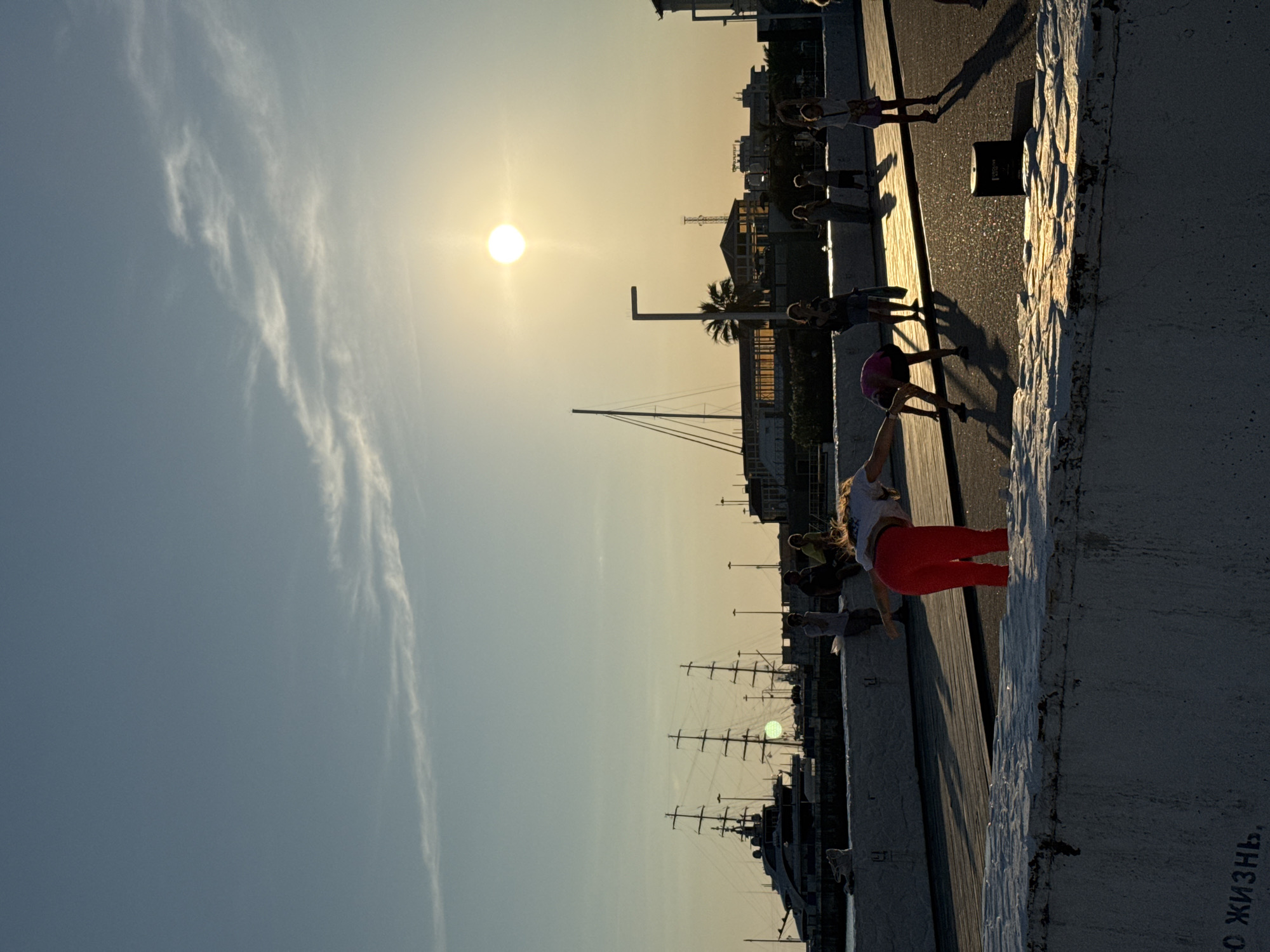 A group of people are engaged in what appears to be an outdoor yoga or exercise class on a pier by the sea. The sun is low in the sky, indicating it is either sunrise or sunset, casting long shadows. The sky is clear with a few wispy clouds. In the background, there are buildings and a palm tree, suggesting a warm climate. Several sailboats are docked in the harbor. One person in the foreground is performing a yoga pose, wearing a white top and red leggings. Another person behind them is also in an active pose. The overall mood seems peaceful and serene, with the golden light of the sun illuminating the scene. There is some graffiti on a concrete wall in the lower right corner of the image, with Russian text that reads "СО ЖИЗНЬ" (SO LIFE).