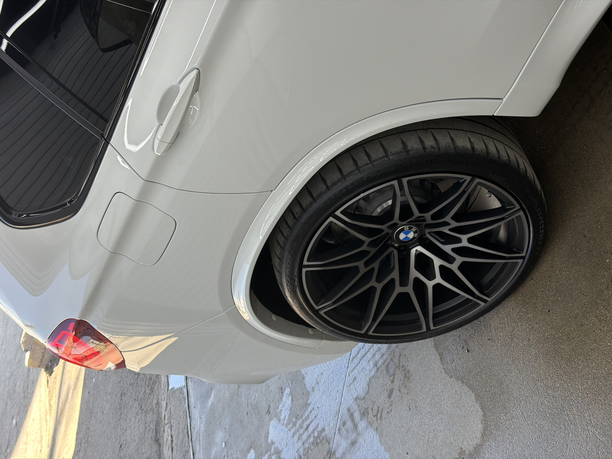 This image displays a close-up of the rear passenger side of a white BMW car, located in Krasnoyarsk, Russia. The vehicle's clean, glossy paintwork and the wet patches on the concrete ground suggest it has been recently washed.
The car features a prominent dark grey, multi-spoke wheel with polished silver accents and a BMW M logo on its hub. A low-profile tire, with "SportContact" text visible on its sidewall, is mounted on the wheel. The car's white body includes a door handle, a subtly flared rear fender, and tinted windows on the passenger side. A fuel cap door is visible on the rear fender, and part of a red taillight can be seen towards the bottom left of the frame.
The setting is an outdoor, paved area, possibly a driveway or car wash bay, under bright daylight conditions, evidenced by strong reflections on the car's surface and the ground. No people are visible in the image.