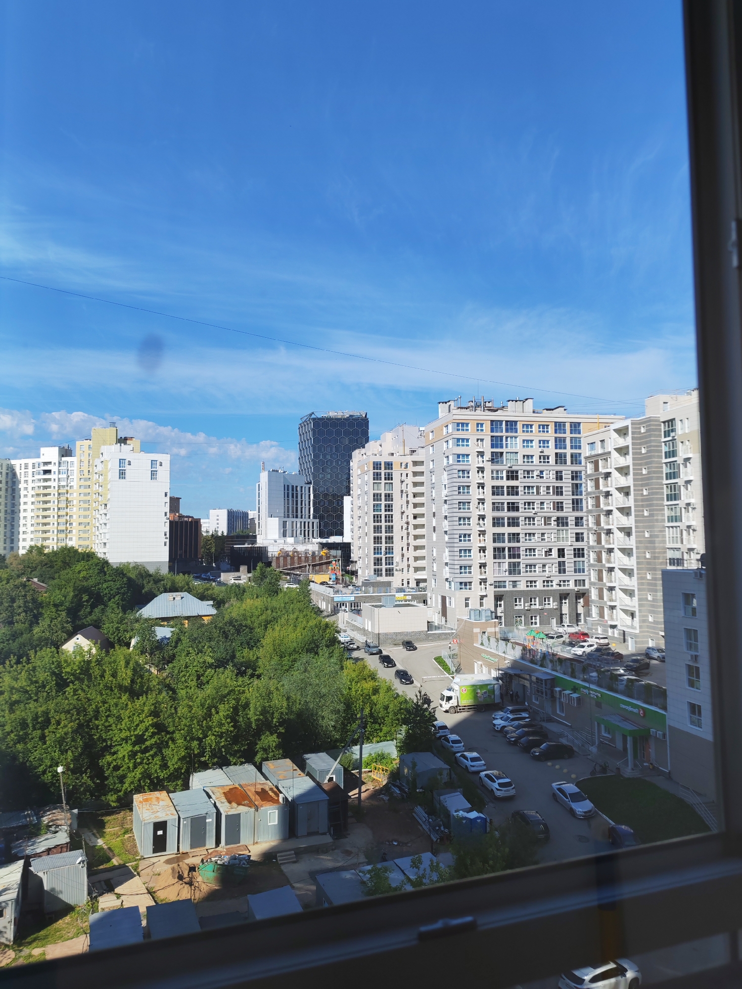 The image shows a daytime view of a modern city with multi-story apartment buildings and some older structures, seen through a window frame. The sky is a clear blue with scattered white cirrus clouds. In the foreground, there are several gray, modular metal buildings, likely storage or temporary structures, with some showing signs of rust. A green dumpster is next to these buildings.
Further back, a street is visible with parked cars and some vehicles in motion. There are also trees and greenery interspersed between the buildings. Dominating the background are several contemporary residential buildings, varying in height and architectural style, some with balconies and numerous windows. One building in the mid-ground has a distinctive hexagonal pattern on its facade. A supermarket with green and white signage is also visible along the street.
Based on visual cues and common architectural styles, this scene is likely located in a residential or mixed-use area of Ufa, Russia, characterized by new construction alongside older industrial or utilitarian structures. No people are clearly identifiable in the image.