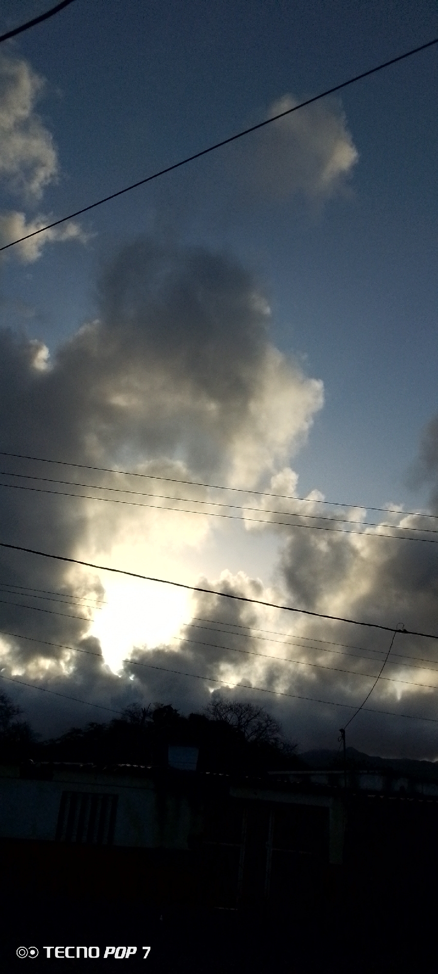 The image captures a view of the sky in Altagracia, Venezuela. Cumulonimbus clouds dominate the scene, partially obscuring the sunlight. A series of overhead electrical wires run across the frame. Below the clouds, silhouetted rooftops and trees are visible. In the lower portion of the image, the corner of a building is also silhouetted. At the very bottom left corner, the phone model used to take the photo, "TECNO POP 7", is visible. The overall tone of the image is dark due to the heavy cloud cover and underexposed foreground elements.