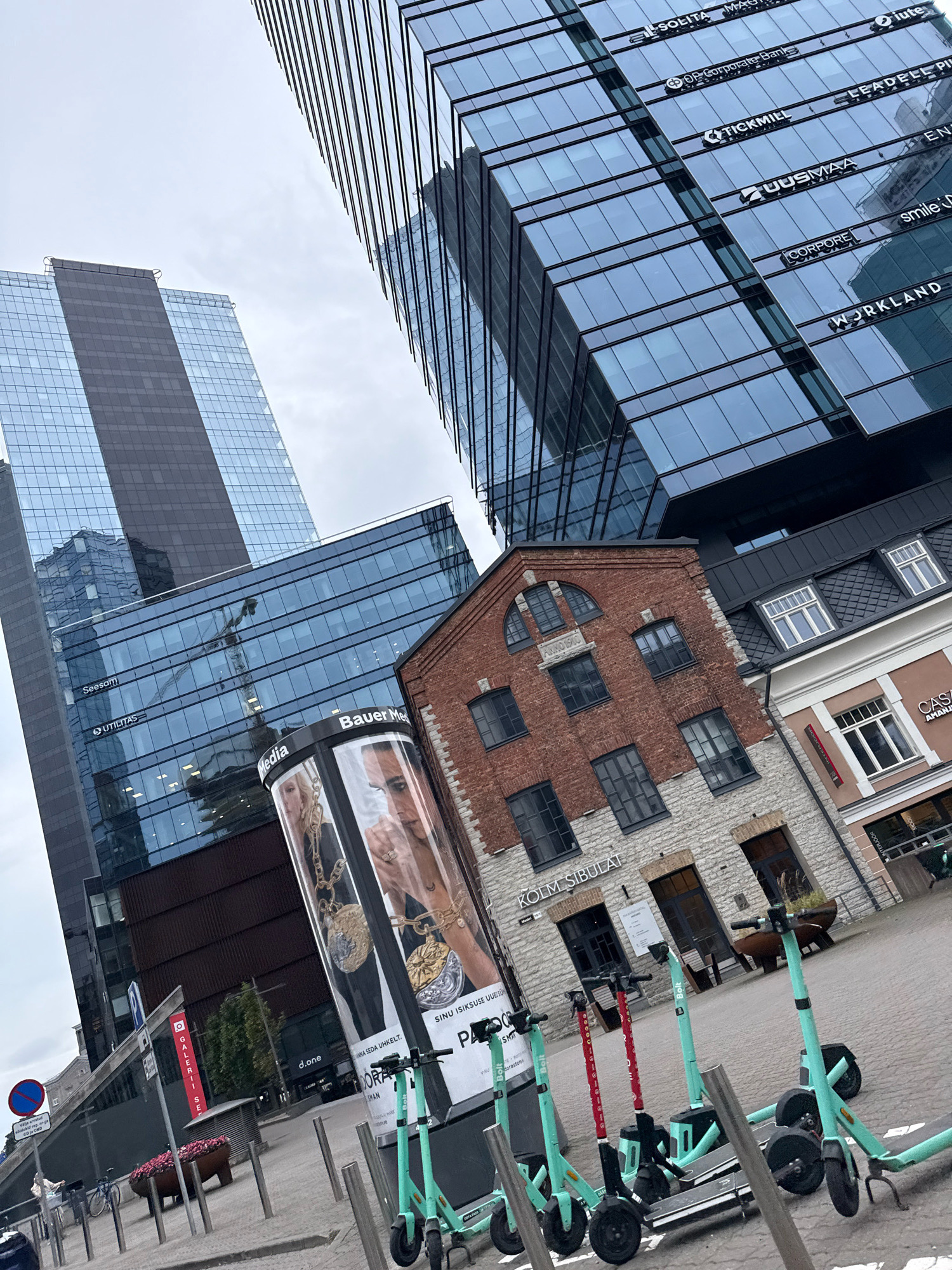 This is an eye-level, wide-angle shot of the modern business district in Tallinn, Estonia, dominated by contemporary glass skyscrapers and a few older brick buildings. In the foreground, several turquoise and red electric scooters are parked. A cylindrical advertising pillar displays a close-up of two people wearing gold jewelry and a text advertisement that reads "SINU ISIKUSE UUEKS". To the left, a blue traffic sign with a red circle and a diagonal line indicates a restriction.
Further back, a series of modern glass buildings rise up, their facades reflecting the cloudy sky. One prominent building features multiple company logos visible on its exterior, including "SOLITA", "MAGNUS", "DF CORPORATE BANK", "TICKMILL", "UUSMAA", "LEADELL PIN", "CORPORE", "WORKLAND", and "smile(d)". Another skyscraper on the left shows a darker, rectangular section on its facade.
Adjacent to the modern structures is a charming, older brick building with arched windows and stone foundations. Above one of its windows, the inscription "ANNO 1910" is visible. To the right of this brick building, another, lighter-colored building with a dark tiled roof and multiple windows is present.
The overall scene suggests a vibrant urban environment where new and old architecture coexist. The presence of electric scooters and advertising pillars points to a lively city center. The weather appears overcast, with a diffuse light. No people are actively engaged in any actions within the frame.
