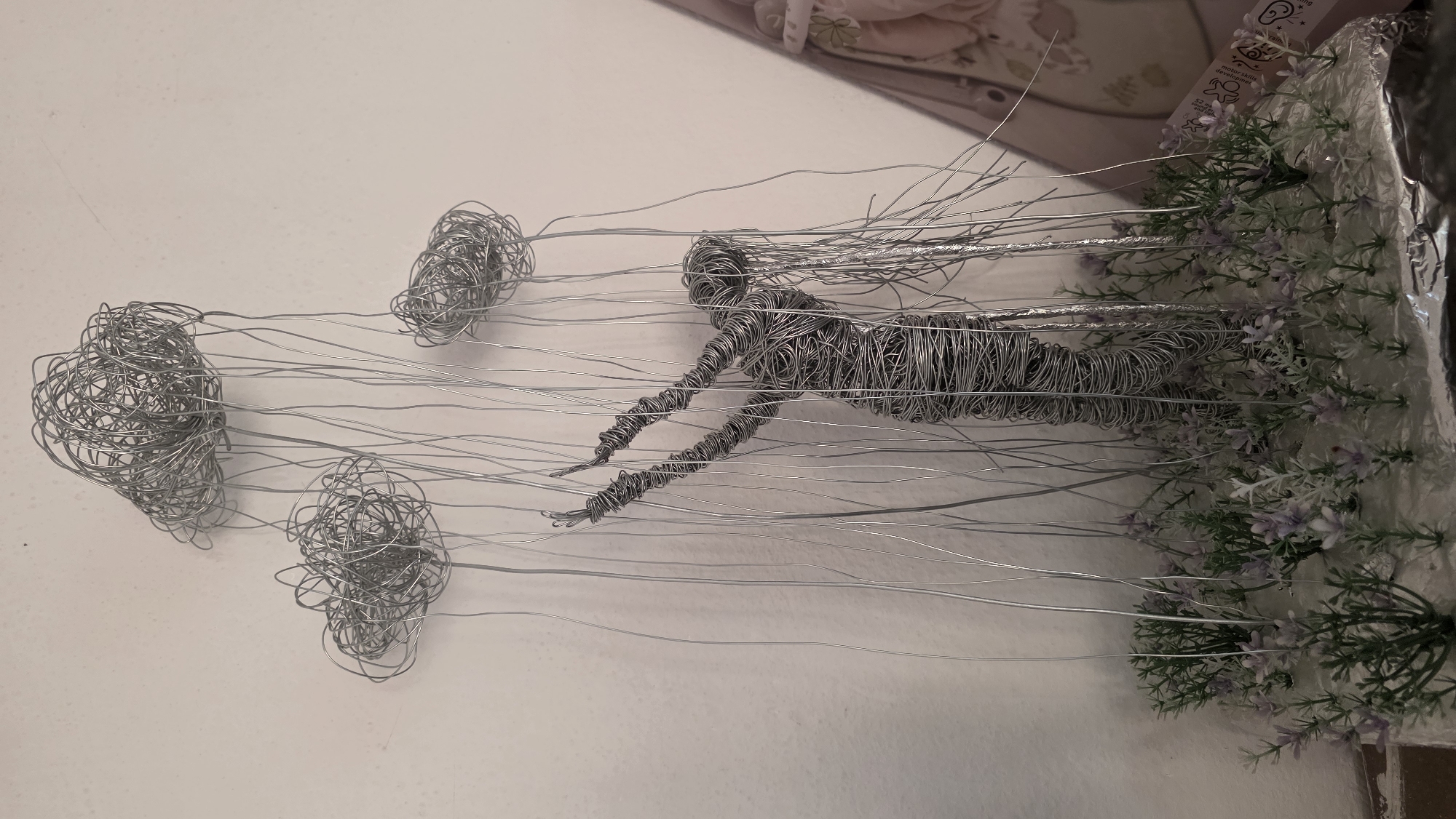 The image shows a wire sculpture of a human figure, seemingly floating horizontally. The figure is constructed from tightly coiled silver wire, forming the body, head, arms, and legs. Attached to one end of the figure are artificial flowers, predominantly green with small purple blooms. At the other end, three clusters of coiled silver wire represent cloud-like formations. Strands of silver wire extend from the figure and the floral arrangement towards the wire clusters, creating a sense of connection and movement. The sculpture is set against a white surface with other objects partially visible in the background. The sculpture is not identifiable with any specific location in Blagoevgrad, Bulgaria.