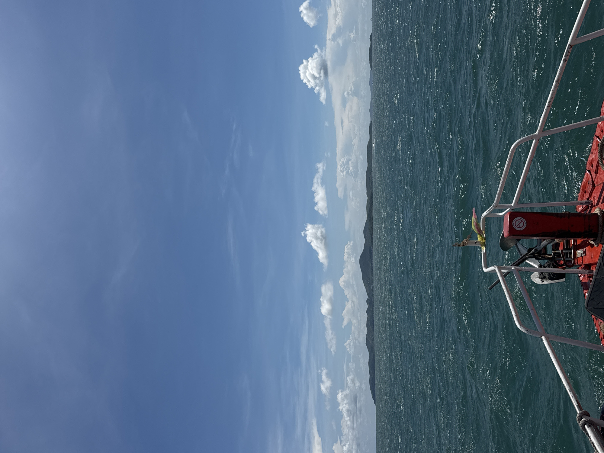A photograph shows a view from a boat out towards the ocean in Pattaya City, Thailand. The image is vertically oriented.
The sky is a clear blue on the left with scattered cumulus clouds running along the horizon line. The ocean occupies the right side of the image, with the boat's railing and a red and white structure visible in the foreground. The water is dark green with sunlight reflecting off the surface. In the distance, land is visible along the horizon line. The scene is bright and sunny.