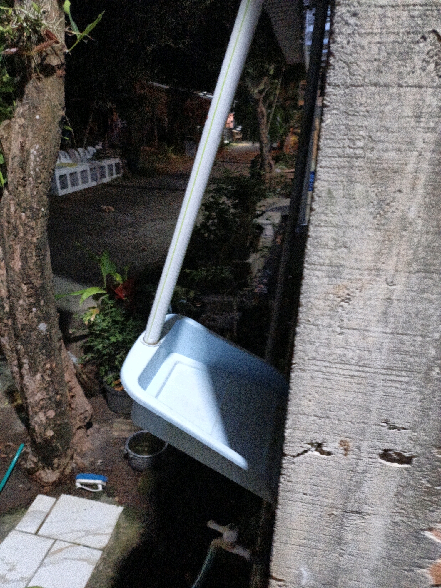 A dustpan with a long handle leans against a rough concrete wall at night. The dustpan is pale blue and appears to be made of plastic. The handle is white with a green stripe. Behind the dustpan, a tree trunk with rough bark is visible on the left. Further back, there are plants in pots, some paving stones, and what appears to be a low concrete structure. The lighting suggests an outdoor, possibly residential, setting in Cikal, Indonesia, as indicated by the request. The scene is dark, with artificial light from an unseen source illuminating the dustpan and the immediate surroundings. No people are present.