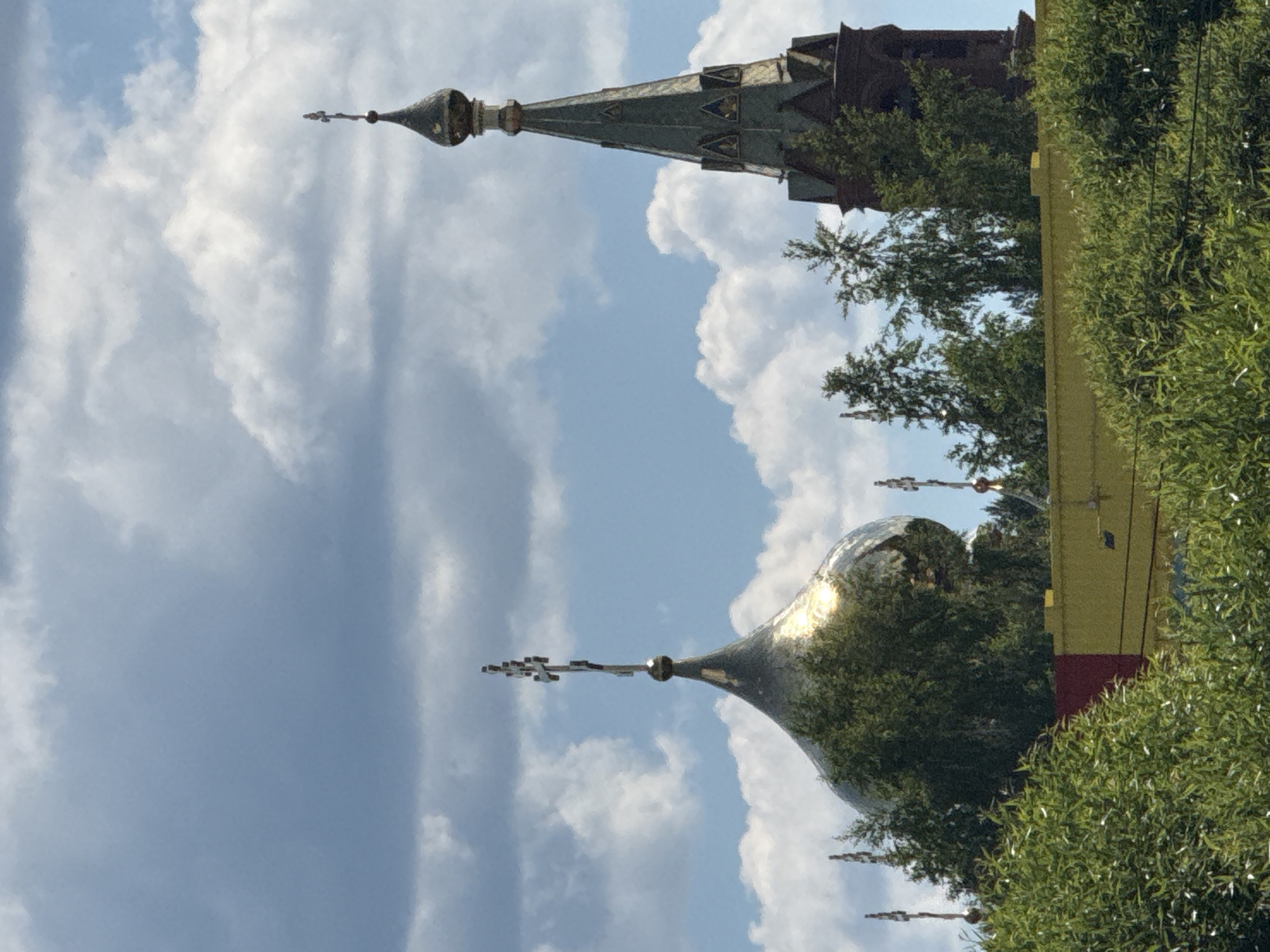 The image captures a view of two church domes and a bell tower peeking through lush green trees under a cloudy sky. The dome on the left is golden and bulbous, topped with a cross. The dome on the right is smaller and darker, also topped with a cross. To the right of these domes is a tall, dark-colored bell tower with arched openings. A yellow building with a red section is visible behind the trees, supporting the structures. The foreground is dominated by dense green foliage.
