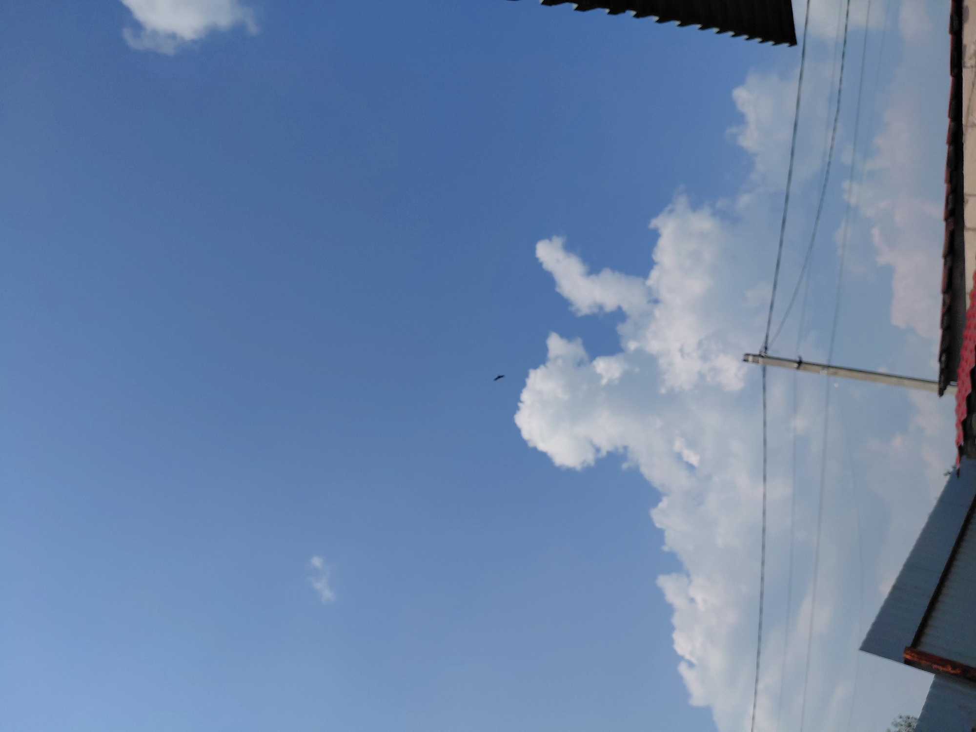 A bright blue sky with scattered white cumulus clouds dominates the upper portion of the image. A single black bird is silhouetted against the sky in the mid-ground. Below the sky, a power line with multiple wires stretches horizontally across the frame. In the lower part of the image, the dark, angled roof of a building and part of another structure with a corrugated metal facade are visible. The scene appears to be an outdoor urban environment, possibly taken from a low angle looking upwards.