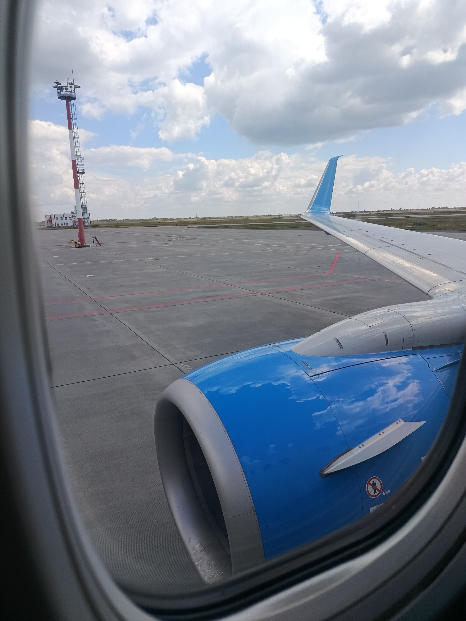 An airplane engine cowling, painted a vibrant blue with a reflective surface showing clouds and sky, is visible from a passenger window. The wing, also partially visible, is white with a blue wingtip. In the background, an airport tarmac stretches out with red markings. A tall, red and white striped control tower stands to the left, with a building at its base. The scene is set on the grounds of the Saburovka Airport in Saburovka, Russia, under a cloudy blue sky. There are no people visible in the image.