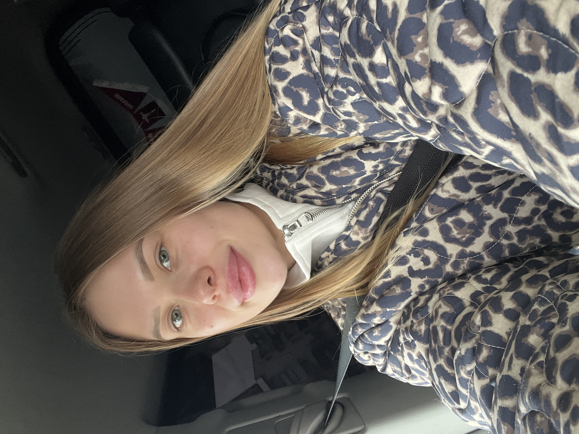 A young woman with blonde hair and fair skin is sitting in what appears to be the passenger seat of a car. She is wearing a leopard-print puffer jacket over a white garment with a zipper. A seatbelt is visible across her torso. The interior of the car is dark. The background shows part of the car's interior and possibly a glimpse of an object with some writing on it. There is no definitive evidence to indicate the city of Chaikovsky, Russia.