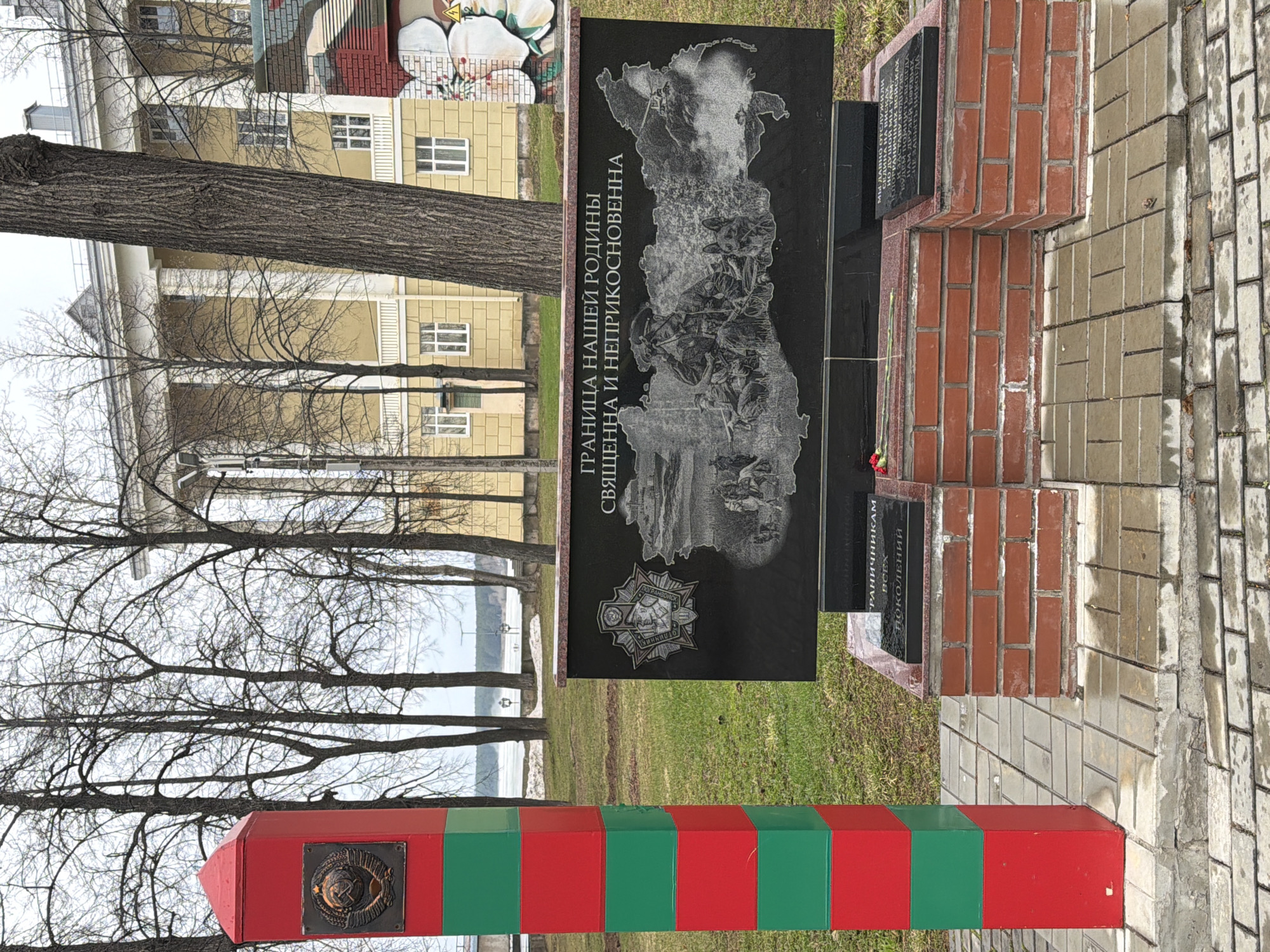 The image captures a memorial dedicated to border guards in Votkinsk, Russia, set in an outdoor public space. In the foreground, a prominent red and green striped pillar, designed like a traditional border post, stands tall. It features a circular bronze plaque with an emblem, likely representing a national or border guard insignia.
Behind the pillar, the central element of the monument is a large, black polished stone slab. This slab is intricately engraved with a map-like outline of a landmass, over which multiple figures of soldiers in combat or on patrol are depicted, symbolizing border defense. Above this scene, a bold inscription in Cyrillic reads: "ГРАНИЦА НАШЕЙ РОДИНЫ СВЯЩЕННА И НЕПРИКОСНОВЕННА," which translates to "THE BORDER OF OUR HOMELAND IS SACRED AND INVULNERABLE." Below the main image, an emblem identifiable as "ПОГРАНВОЙСКА" (Border Troops) is also engraved.
The black slab is mounted on a base constructed from reddish-brown bricks. On the right side of this brick base, a smaller black plaque bears the dedication: "ПОГРАНИЧНИКАМ ВСЕХ ПОКОЛЕНИЙ," meaning "TO BORDER GUARDS OF ALL GENERATIONS." Another partially visible plaque next to it contains further dedicatory text. Red flowers, possibly carnations, are placed at the base of the monument, indicating remembrance or a recent tribute.
The surrounding environment includes several bare trees, their branches reaching towards an overcast sky. Through these trees, a glimpse of a large body of water, potentially a lake or river, can be seen in the distance, possibly with ice reflecting the dull light. A large, light-yellow building with numerous windows is visible in the background. The ground consists of green grass around the monument and a paved walkway of light-colored interlocking bricks. The scene conveys a solemn and respectful atmosphere, likely a memorial to those who served to protect the nation's borders.