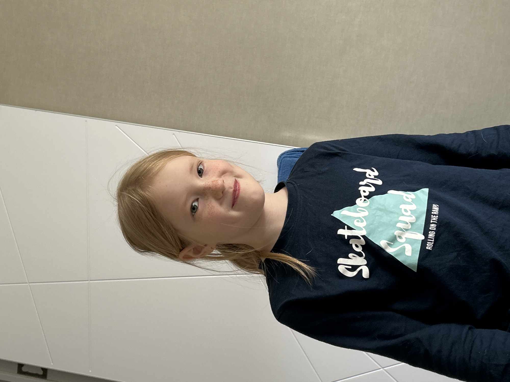 An image, rotated 90 degrees clockwise, features a young girl with blonde hair and numerous freckles on her nose and cheeks. She has light-colored eyes and is smiling gently while looking towards the camera. She is wearing a navy blue long-sleeved t-shirt. On the front of her shirt, there is text that reads "Skateboard Squad" in white script, with "Squad" prominently displayed on a light blue triangular graphic. Below this, in smaller white letters, it says "ROLLING ON THE RAMPS." The background consists of plain, light-colored walls, possibly tiled, suggesting an indoor setting. The girl appears happy and comfortable. The location context provided for this image is Chaikovsky, Russia.
