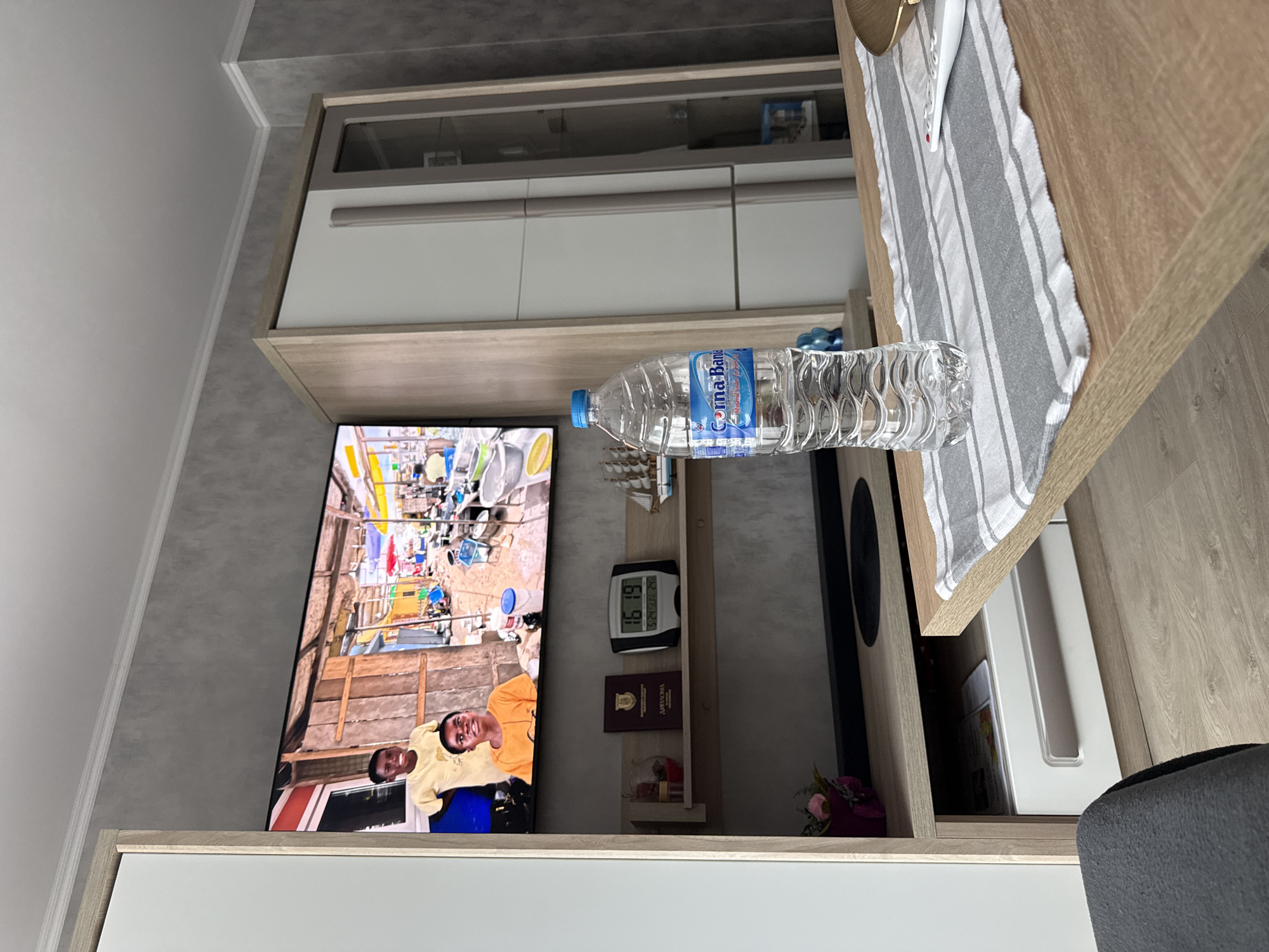 The image depicts an interior scene in Lovech, Bulgaria. A plastic bottle of "Gorna Bania" mineral water is placed on a wooden table covered with a striped cloth. In the background, a television displays a scene involving two people. Under the television, a shelf unit features a digital clock showing 16:19, a diploma, and other small objects. Above the TV and shelves are cabinets with white fronts and wooden frames. The room has gray walls, and part of a gray chair is visible in the lower right corner.