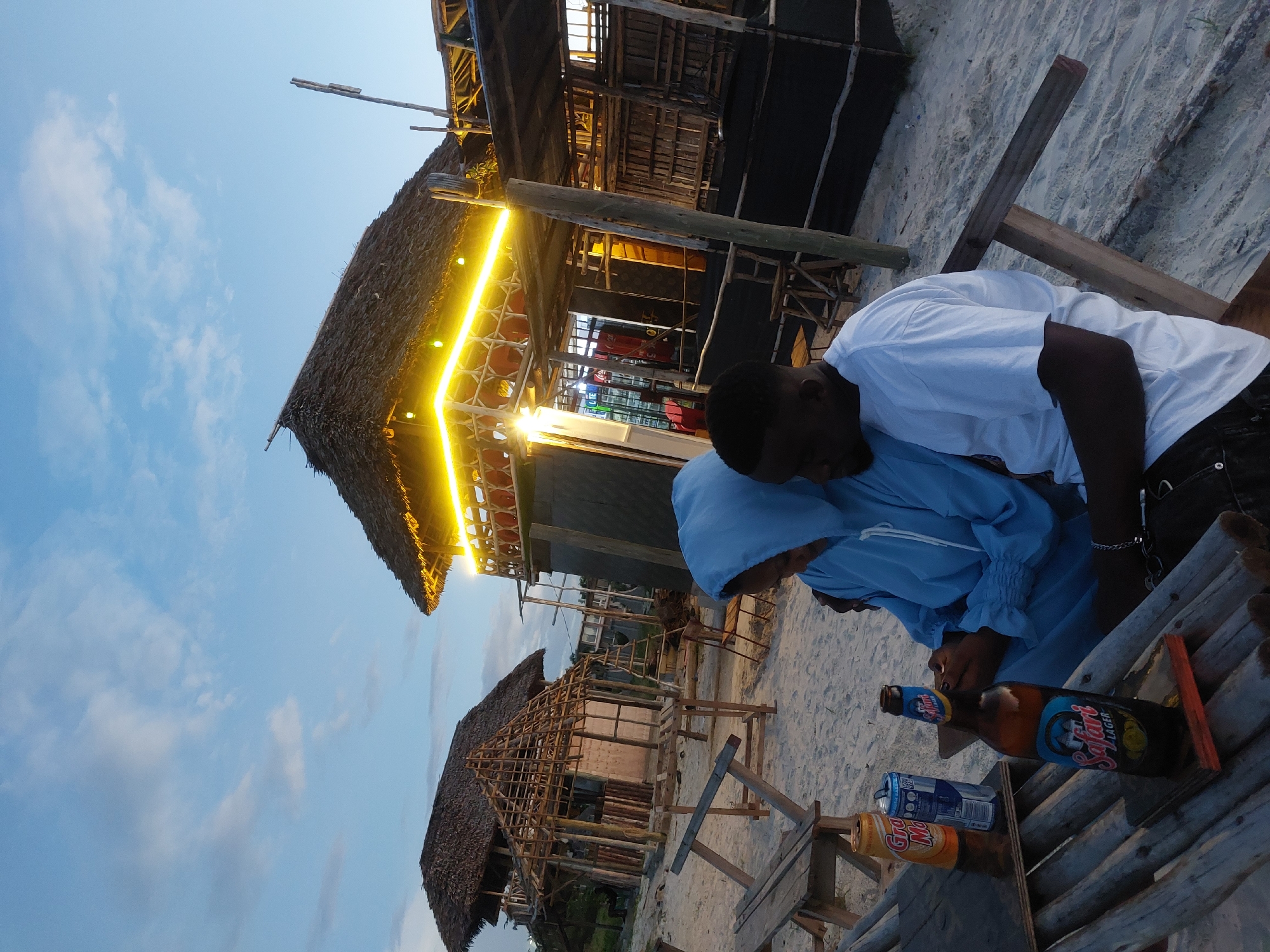 Two people, a man in a white t-shirt and a person in a blue jacket, are sitting at a wooden table on a sandy beach. They appear to be looking at or interacting with something on the table. On the table, there are several beverage containers, including a Safari Lager beer bottle and cans of soda. In the background, there are buildings with thatched roofs, one of which is illuminated with yellow lights. The scene is set in Dar es-Salaam, Tanzania, as evidenced by the Safari Lager beer, which is a local Tanzanian brand. The sky is a gradient of blue and the light suggests it is either dusk or dawn.