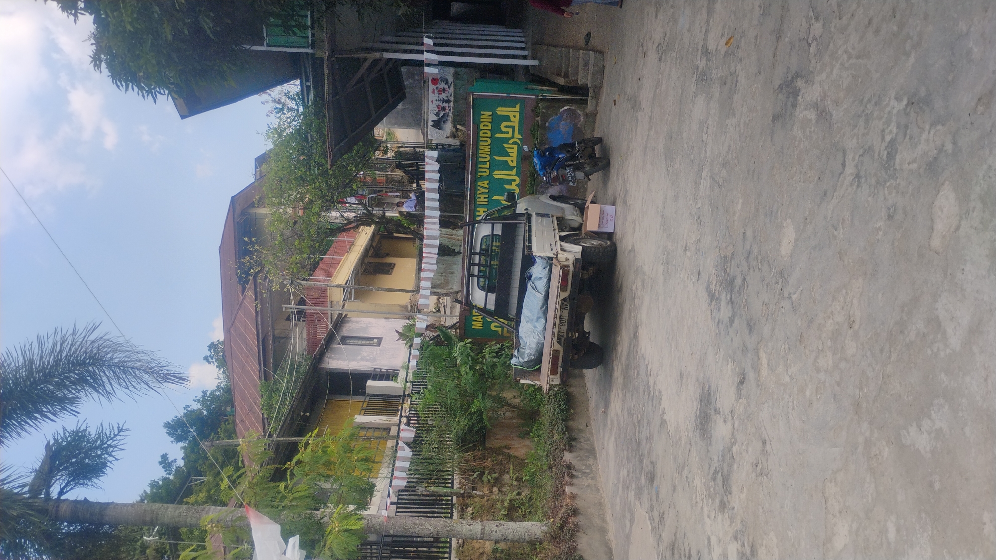 In Samarinda, Indonesia, a light-colored pickup truck is parked on a concrete surface. The truck bed is covered with a light blue tarp, obscuring its contents. To the right of the truck, a motorcycle is parked. In front of the truck, a cardboard box sits on the ground. A large green sign with Arabic script and text in Indonesian is visible behind the truck. The background features buildings with tiled roofs and trees, suggesting an outdoor setting. The sky is partly cloudy.