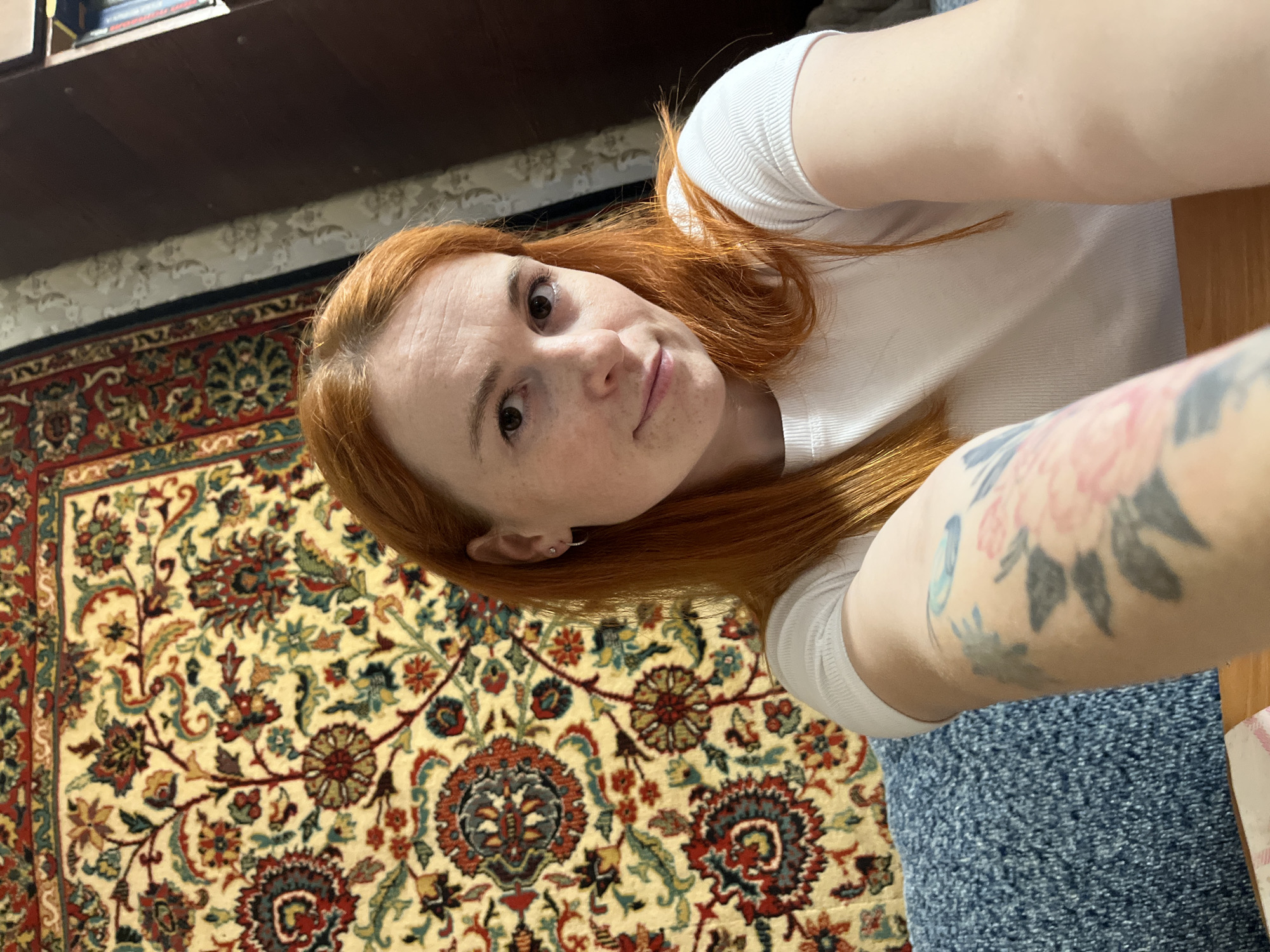 An adult female with reddish-orange hair and fair skin is taking a selfie in an indoor setting. She is wearing a white t-shirt and has a tattoo on her left arm which appears to be floral. She is sitting in front of a tapestry with a red, yellow, and blue floral pattern. To her right, a wooden bookshelf is visible, containing books. The woman is looking directly at the camera with a slight smile. The background suggests a home environment, potentially in Chaikovsky, Russia, based on the provided context.