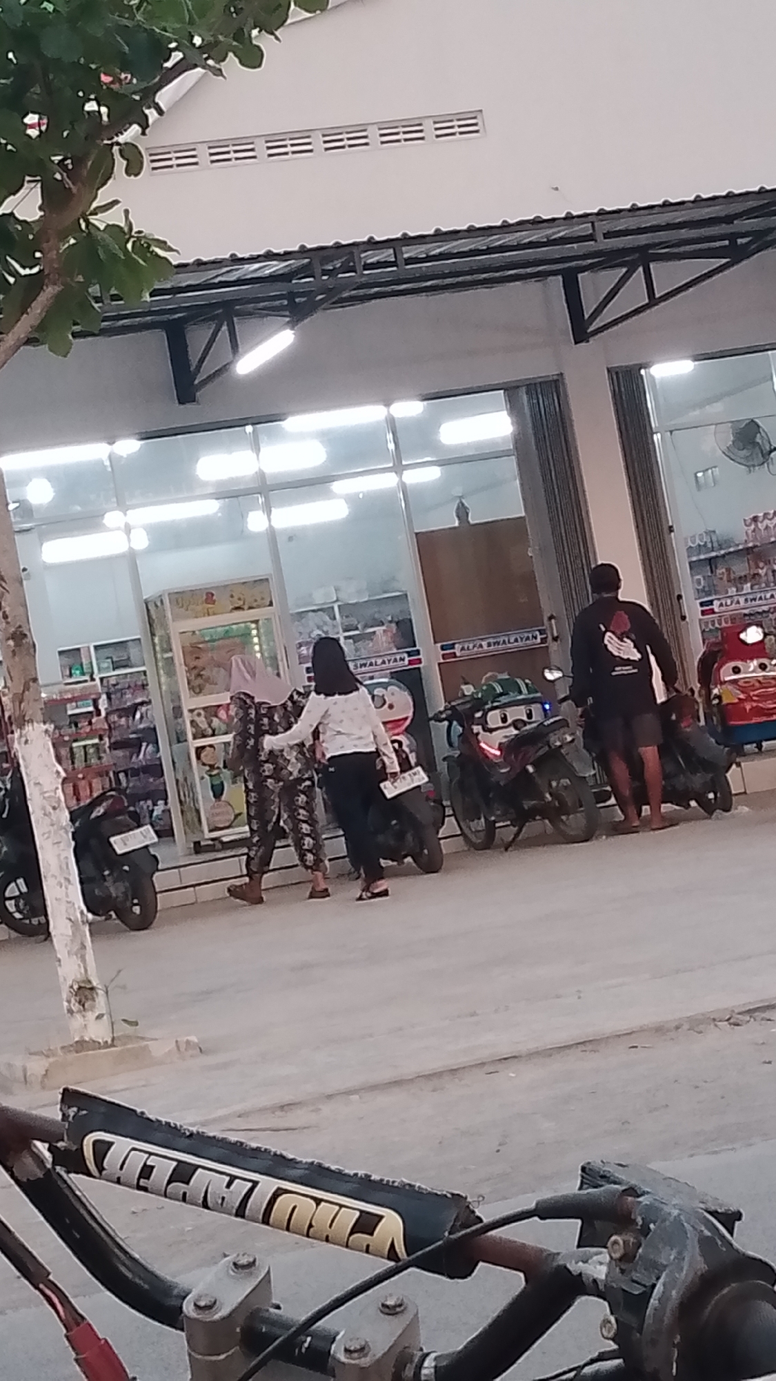 A medium shot captures a street scene in front of an Alfa Swatayan convenience store in Karanglangu, Indonesia. Two women and a man are present. One woman, wearing a patterned outfit and a headscarf, is interacting with another woman dressed in a white long-sleeved shirt and black pants. The man, in a black t-shirt and shorts, stands nearby. Several motorcycles are parked in front of the store. The store has a glass facade, revealing shelves of products inside. The overall lighting suggests it is late afternoon or early evening. The foreground of the image is dominated by the handlebars of a motorcycle, showing a "Pro Taper" grip.