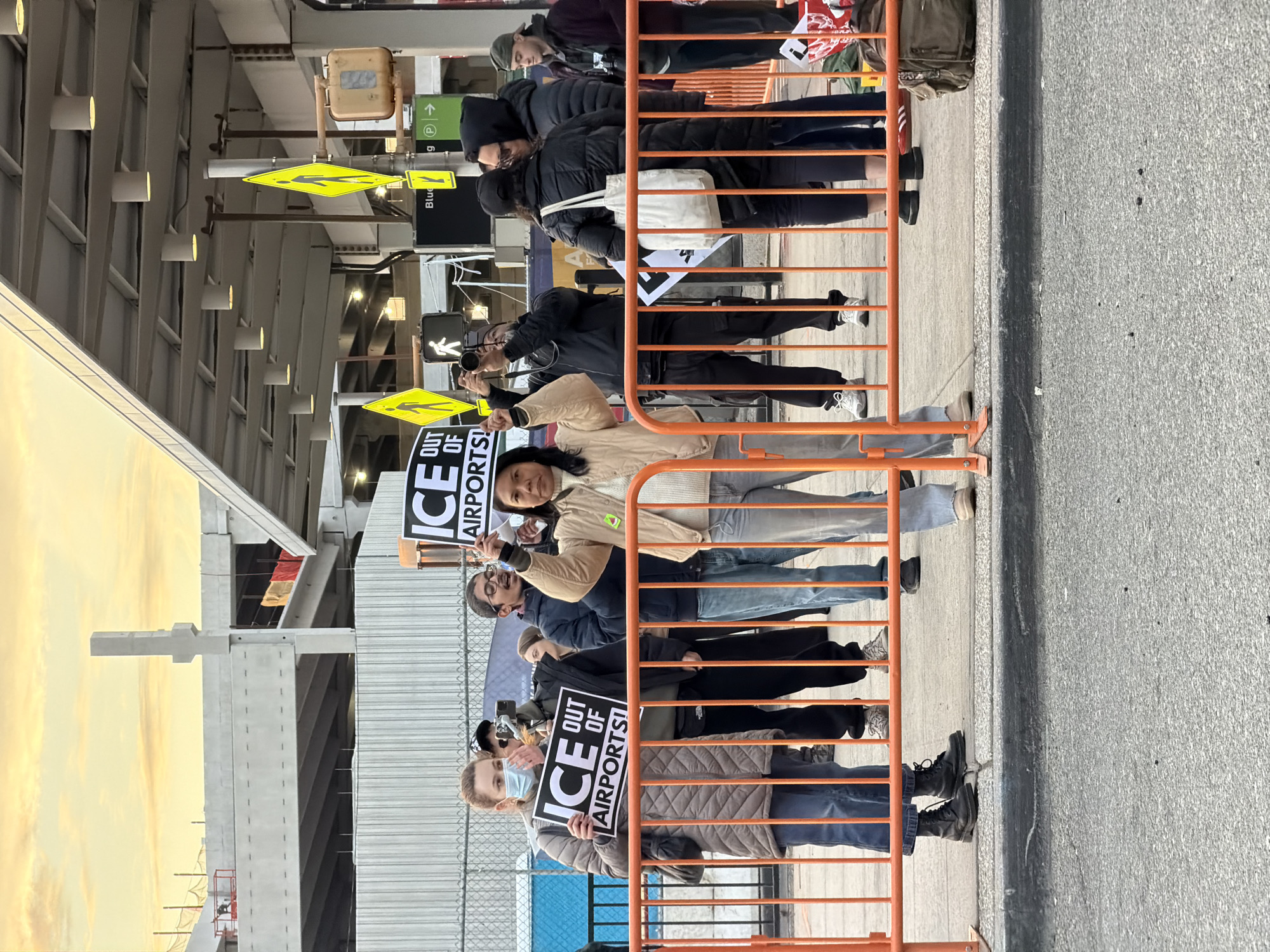 The scene depicts an outdoor protest occurring at LaGuardia Airport (LGA) in New York, United States of America. The time is late afternoon, characterized by a golden-orange sky on the horizon. Approximately 15 individuals are gathered behind orange metal crowd control barriers.
Two female protestors are prominently featured. One on the left, wearing a brown puffer coat, blue jeans, and a light blue medical mask, holds a black and white sign reading "ICE OUT OF AIRPORTS!". Another female protestor in the center, wearing a cream sweater and a beige jacket, holds an identical sign above her head. Multiple other individuals are present, some observing, others actively photographing the event with cameras; one person in dark clothing on the left holds a camera to their eye, and another in the center-right aims a camera forward.
The background consists of modern airport infrastructure, including an elevated concrete structure in a state of ongoing construction, an overhead canopy with recessed lighting, and various airport signage. Visible signs include multiple yellow diamond-shaped pedestrian crossing signs, a "Blue" sign, and a green directional arrow sign. The ground in the immediate foreground is asphalt, transitioning to concrete pavement behind the barriers. A brown backpack rests on the ground to the far right.