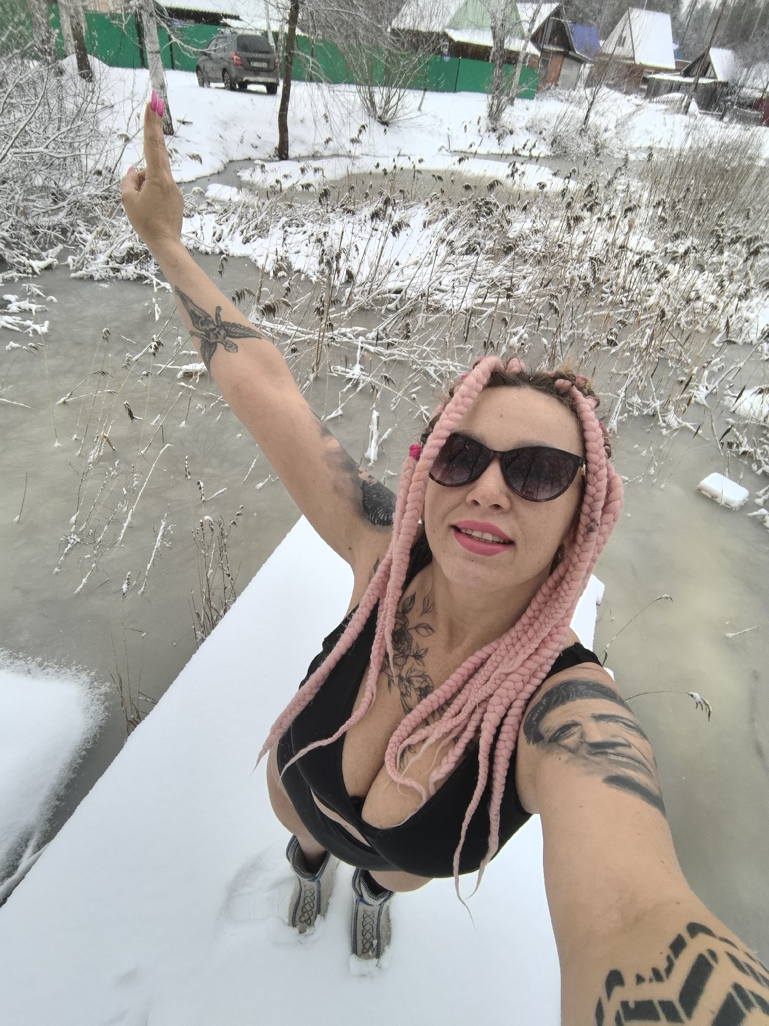 A woman takes a selfie on a snow-covered dock, with a partially frozen body of water behind her.
**People:**
* A light-skinned woman with pink braided hair, wearing sunglasses, a black top, and boots. Her left arm is raised with the index finger pointing upward. She has multiple tattoos on her arms and chest.
**Environment:**
* Winter scene, likely in a rural area. Snow covers the ground and dock.
* A partially frozen body of water behind the dock with snowy reeds.
* In the background, trees, houses, and a parked car are visible.
**Objects:**
* Snow-covered wooden dock
* Boots
* Sunglasses
* Mobile phone
* Black SUV vehicle in the background
* Pink Braids
* Frozen water
* Reeds
**Actions:**
* The woman is taking a selfie, holding a phone in her right hand.
* She is standing on the dock in the snow.
* She is pointing upward with her left index finger.
**Location:**
* Likely Noviy, Russia, based on the visual characteristics of the environment (architecture, vegetation).