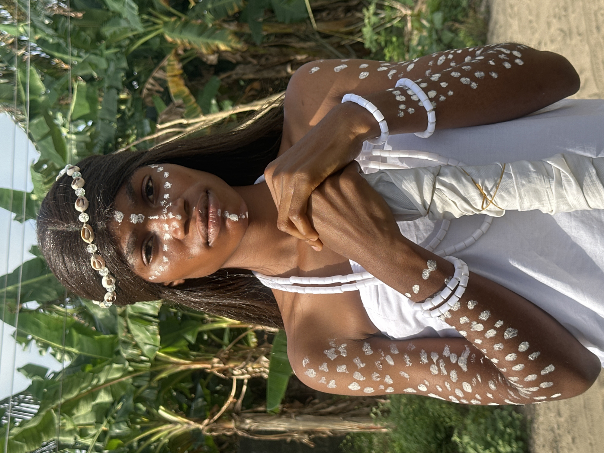 A young Black woman stands facing the camera, adorned in white body paint and white beaded jewelry, with a shell-adorned headband. She wears a white garment that covers her chest and shoulders. Her arms, chest, and face are decorated with white markings, some resembling dots and others more abstract patterns. She clutches a white-wrapped object with both hands, holding it in front of her chest. The background features lush green banana trees and foliage, suggesting an outdoor, possibly tropical environment. The lighting indicates it is daytime, with sunlight illuminating the scene. The overall impression is one of cultural or spiritual attire. This image was taken in Badagry, Nigeria.