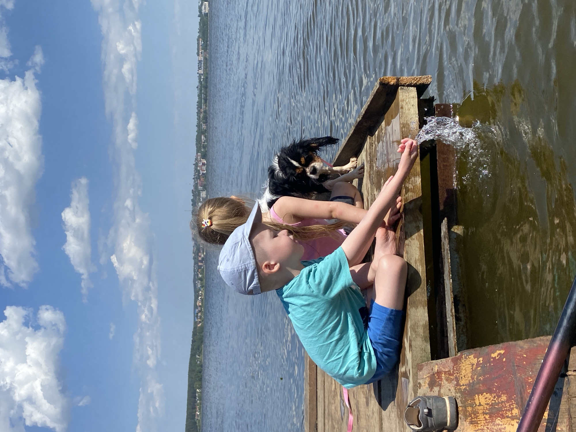 Two children, a girl and a boy, sit on a wooden dock with a dog, extending their hands into the water. The boy wears a blue shirt, blue shorts, and a light blue hat. The girl has blonde hair tied back with a hair tie and wears a pink top. A black and white dog stands behind them. A shoe sits at the edge of the dock, partially obscured. Water splashes up as they interact with it. In the background, a body of water extends to the horizon, where buildings are visible, suggesting the location is Votkinsk, Russia. The sky is blue with scattered clouds.